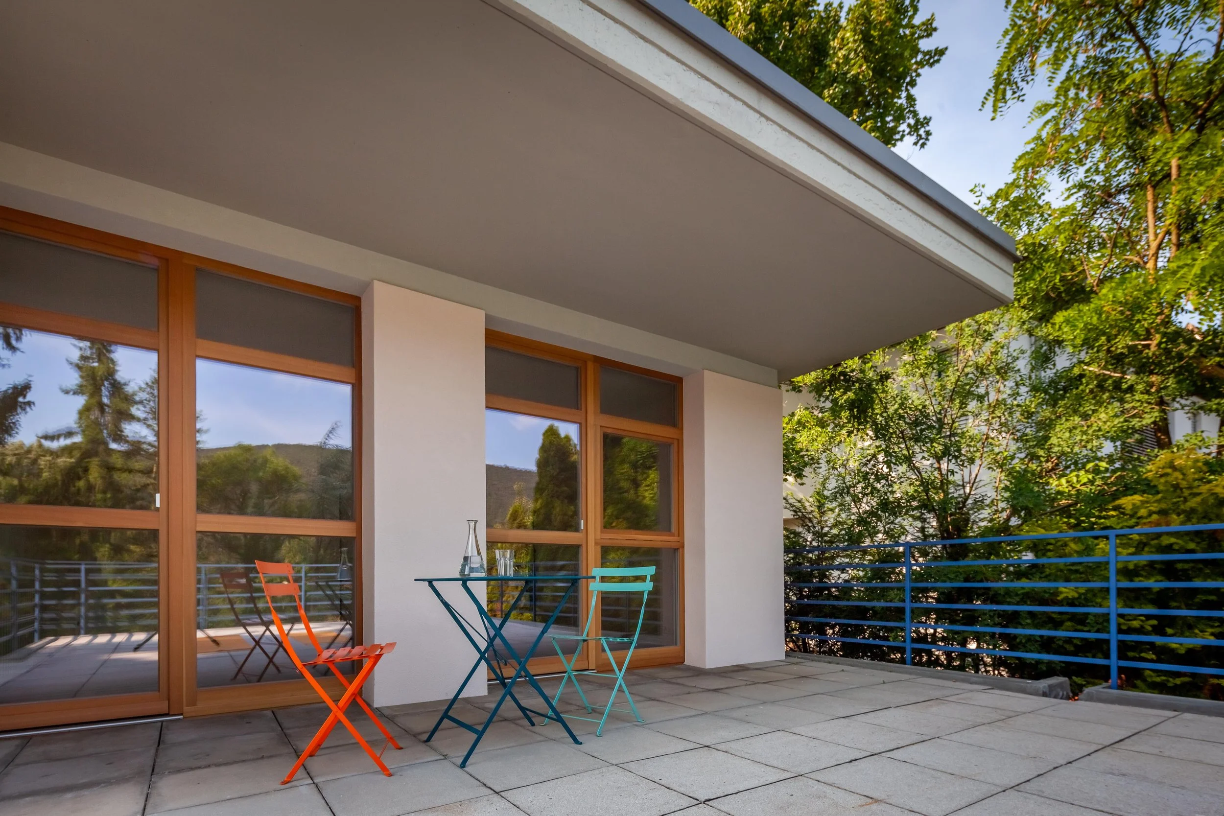 Balkon mit drei bunten Stühlen und einem kleinen Tisch, umliegendes Grün, moderne Fenster und blaue Geländer.