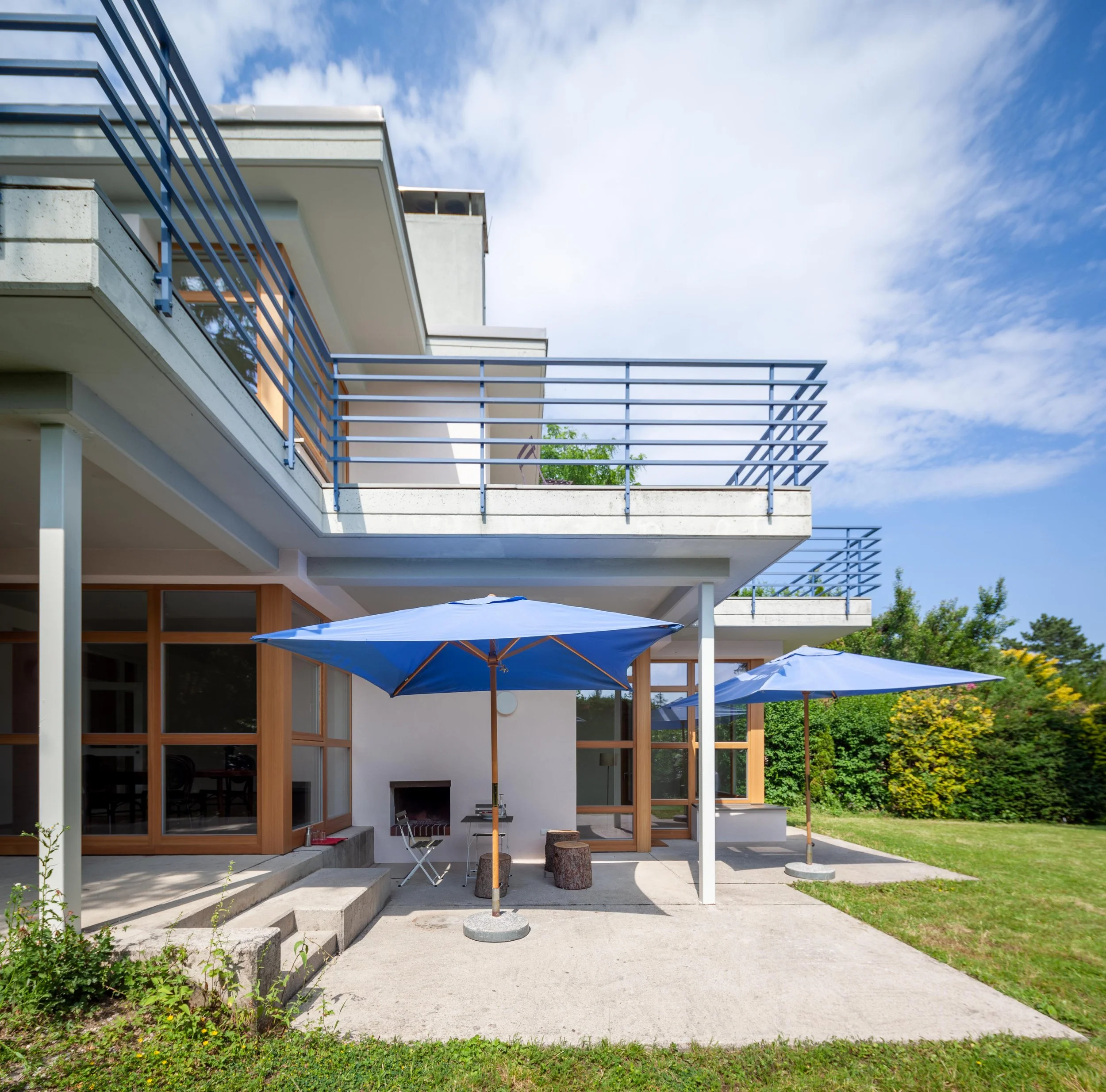 Moderner Hausgarten mit Terrasse, zwei blaue Sonnenschirme, Holzmöbeln, Stümpfen und Garten mit Sträuchern und Bäumen, blauer Himmel mit wenigen Wolken.