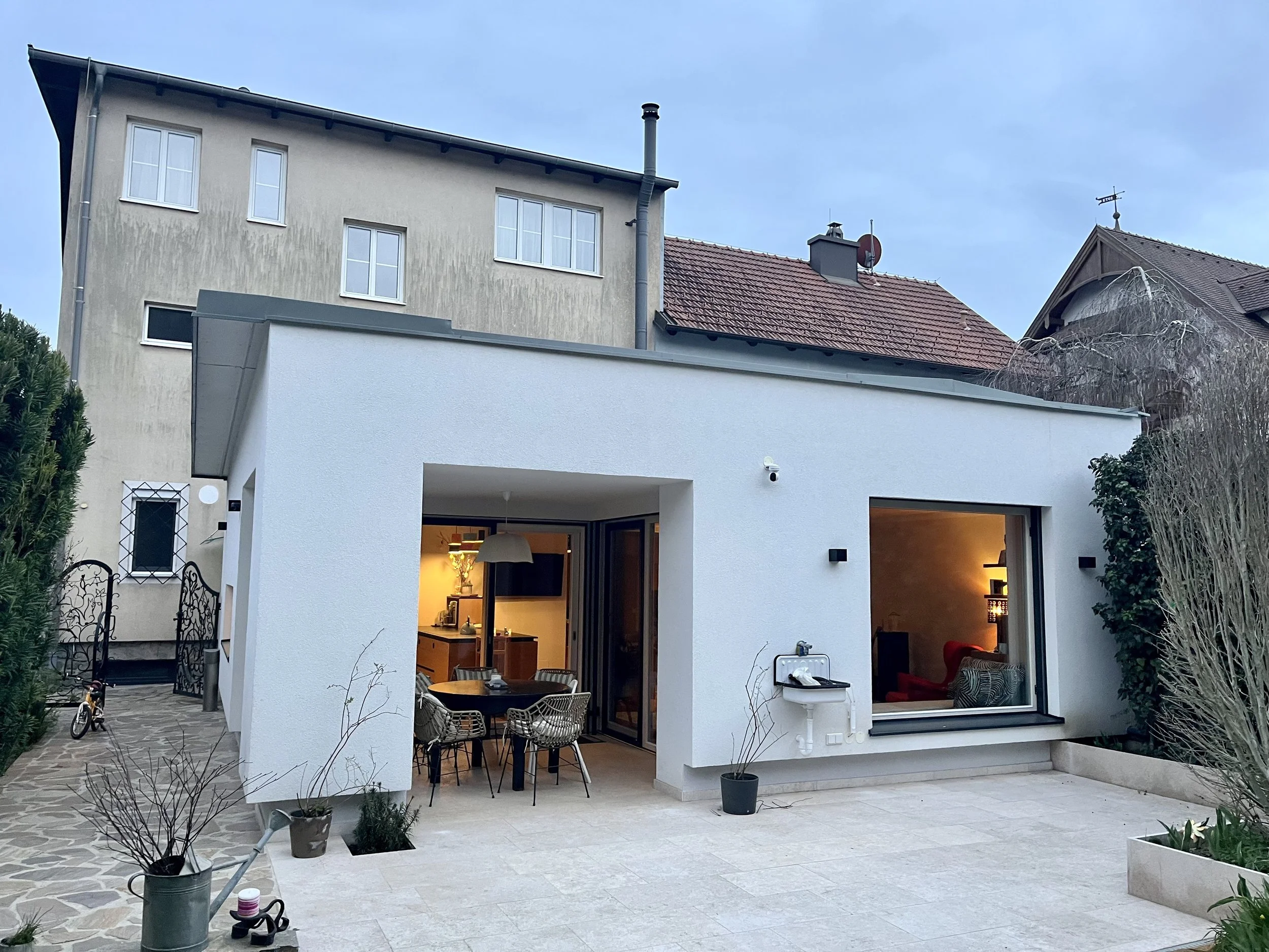 Modernes weißes Haus mit offener Terrasse, beleuchtetem Innenraum, Garten mit Pflanzen, Spielzeug Fahrrad, auf dem Hinterhof, am Abend.
