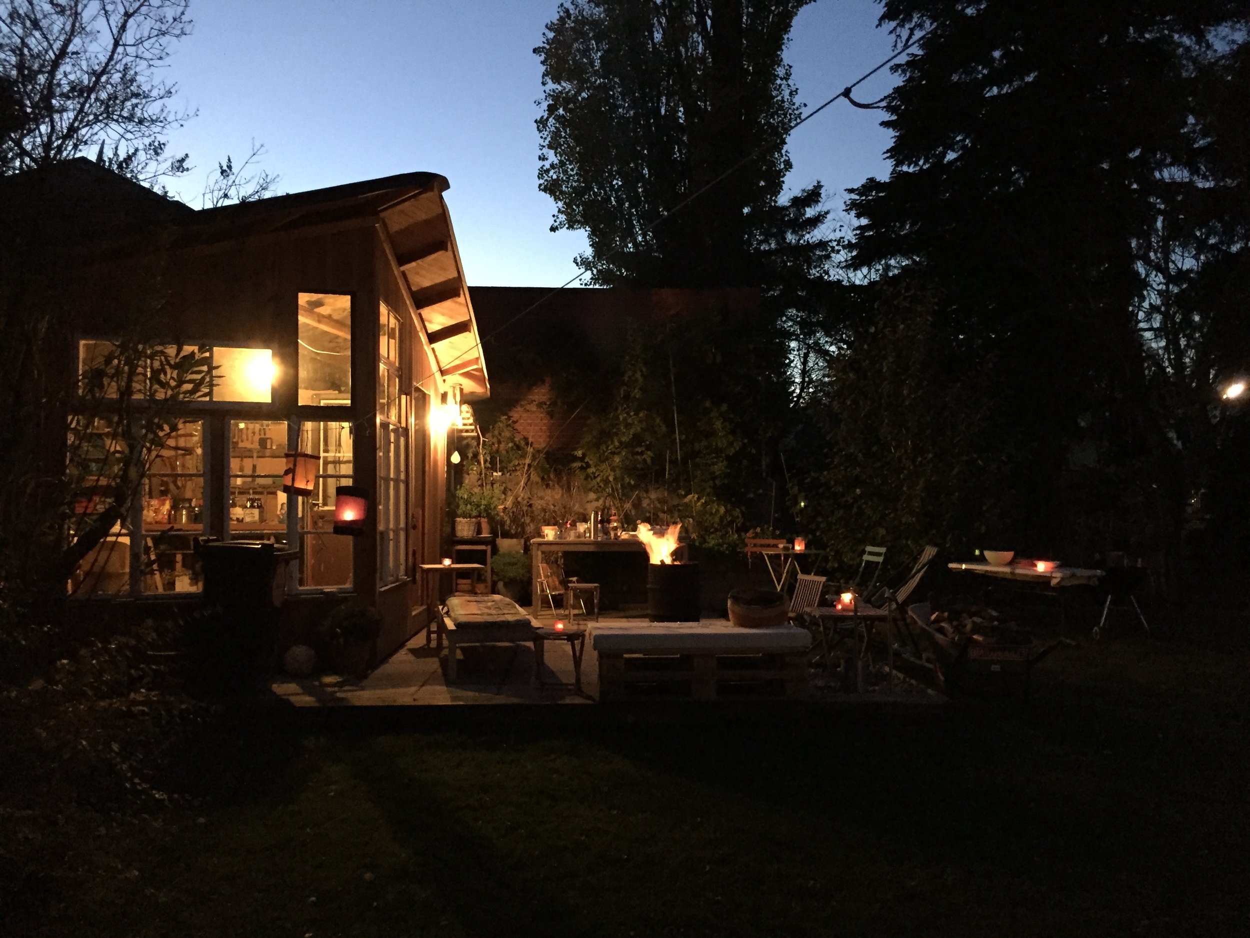 Garten bei Dämmerung mit gemütlicher Beleuchtung, einem Feuer im Feuerkorb, Gartenmöbeln und Lichterketten, Blick auf einen Glashäuschen oder eine Hütte.