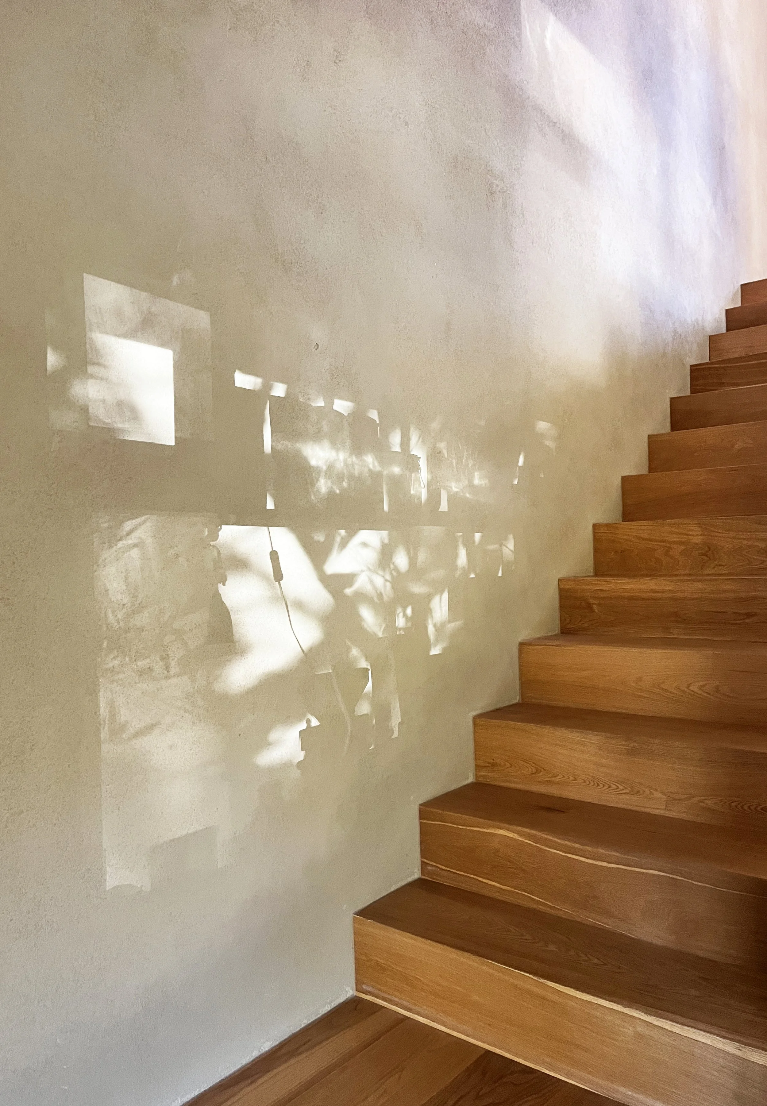 Schatten einer Tischlampe wirft geometrische Muster an eine weiße Wand neben einer Holztreppe.