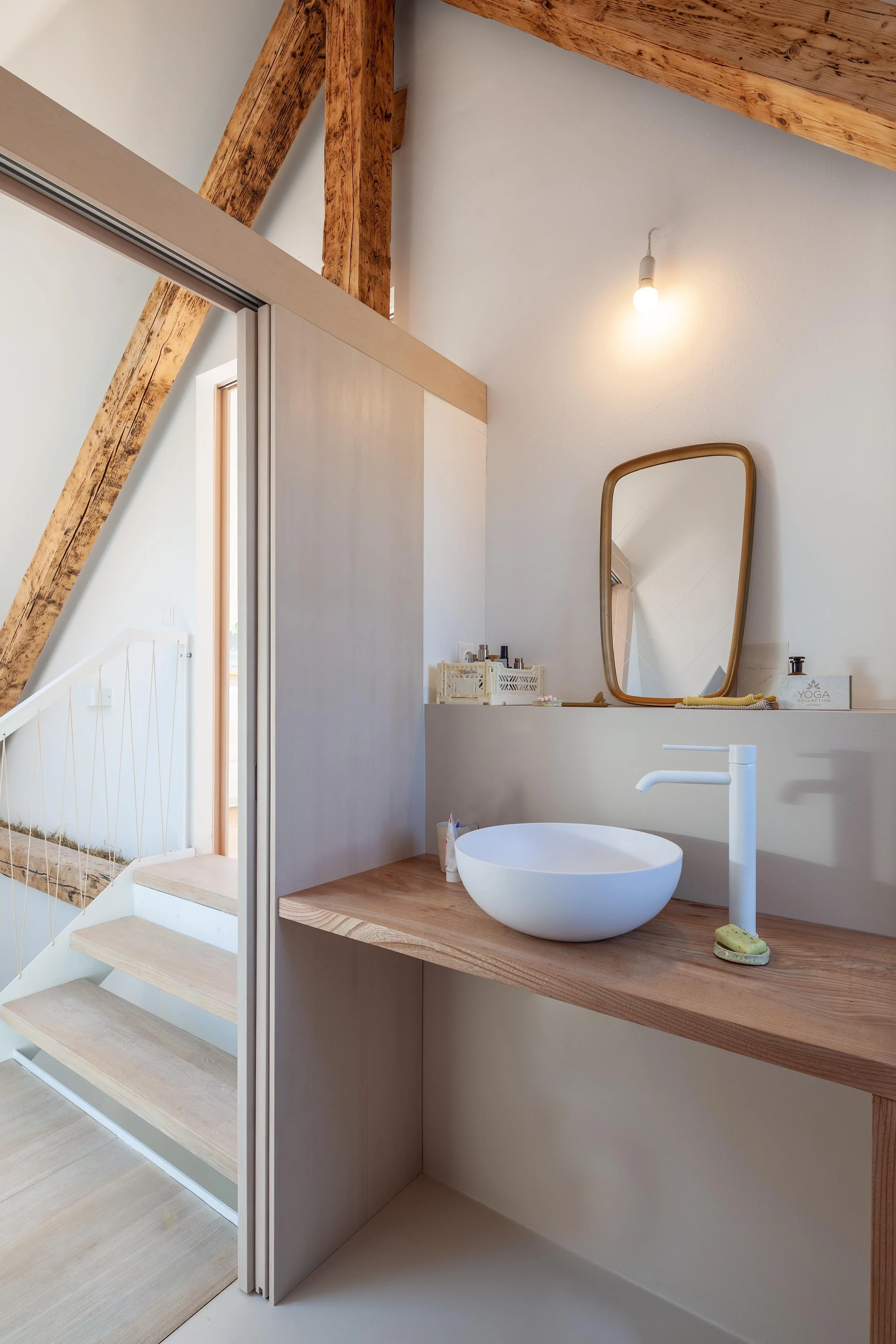 Modernes Badezimmer mit Holz-Waschbecken, ovalem Spiegel, weißen Keramikwaschbecken, einem Handtuch, Kosmetikartikeln und Holzträgern. Helles, minimalistisches Design mit sichtbaren Holzbalken und einer Glastür, die in den Treppenaufgang führt.