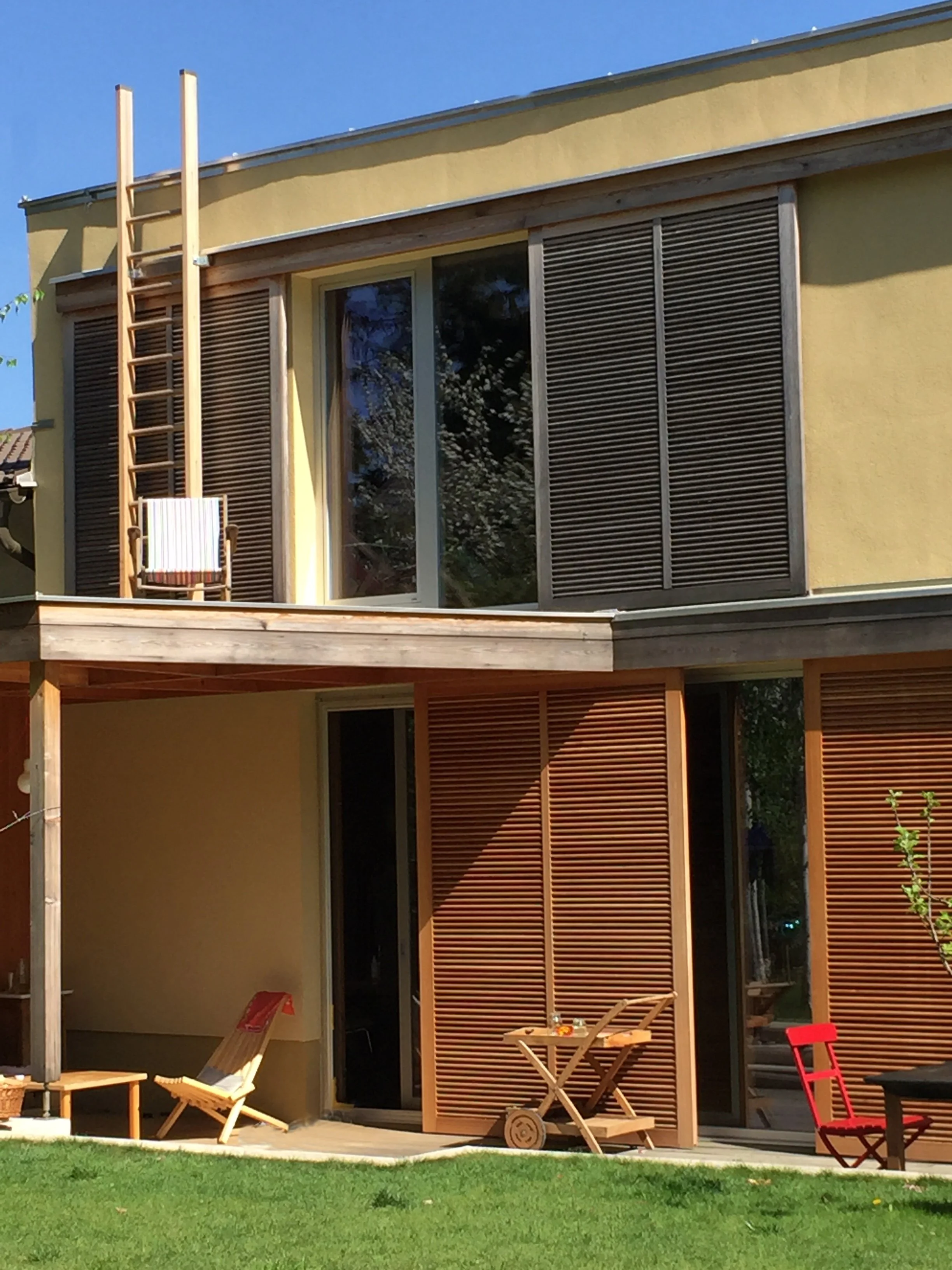 Zweistöckiges Haus mit gelber Fassade, Holzjalousien und Balkon, Garten mit Stühlen und Tisch, sonniger Tag, blauer Himmel.