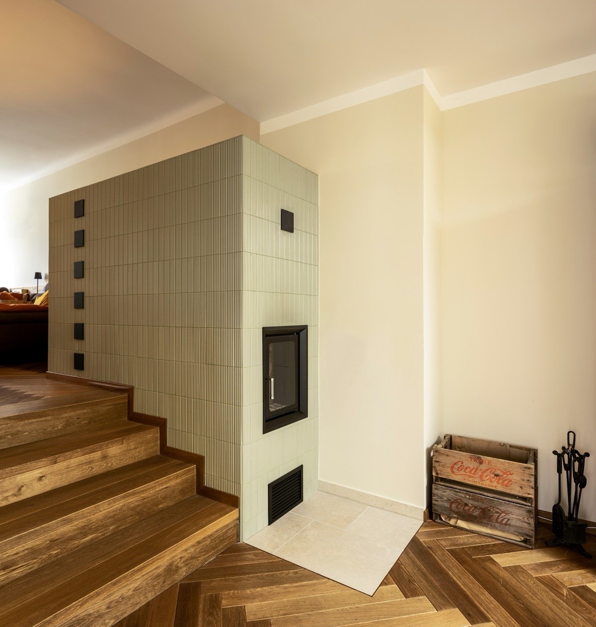 Ein moderner Kamin in einem Wohnzimmer neben einer Treppe aus Holz, mit einem Holz- und Coca-Cola-Box im Ecke, die auch Werkzeug oder Feuerzubehör enthält.