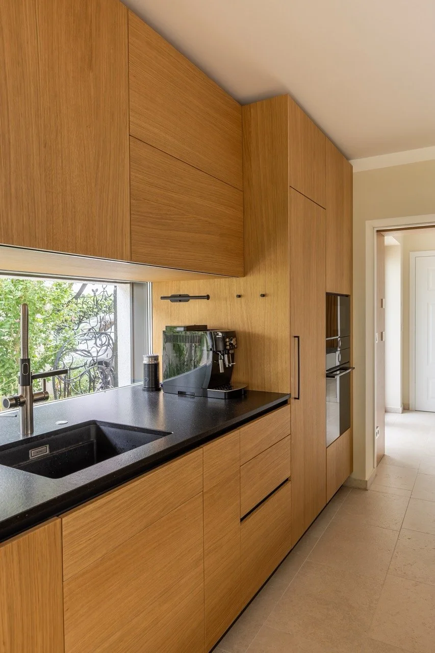 Moderne Küche mit Holzschränken, schwarzer Theke und Kaffeemaschine neben Fenster mit Blick auf grüne Bäume.