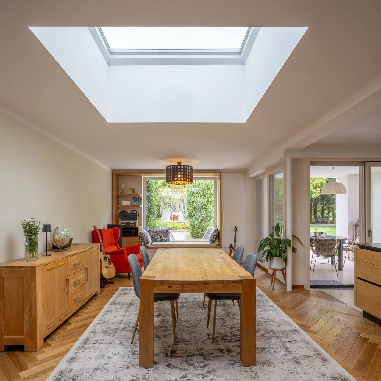 Helle, modernisierte Wohnzimmer mit Essbereich, Holzfußboden, großer Dachfenster, Wohnzimmer mit großem Fenster zur Garten, Möblierung mit hellen Holzschränken, roter Sessel, Gitarre, Pflanzen, Balkon mit Balkonmöbeln draußen.