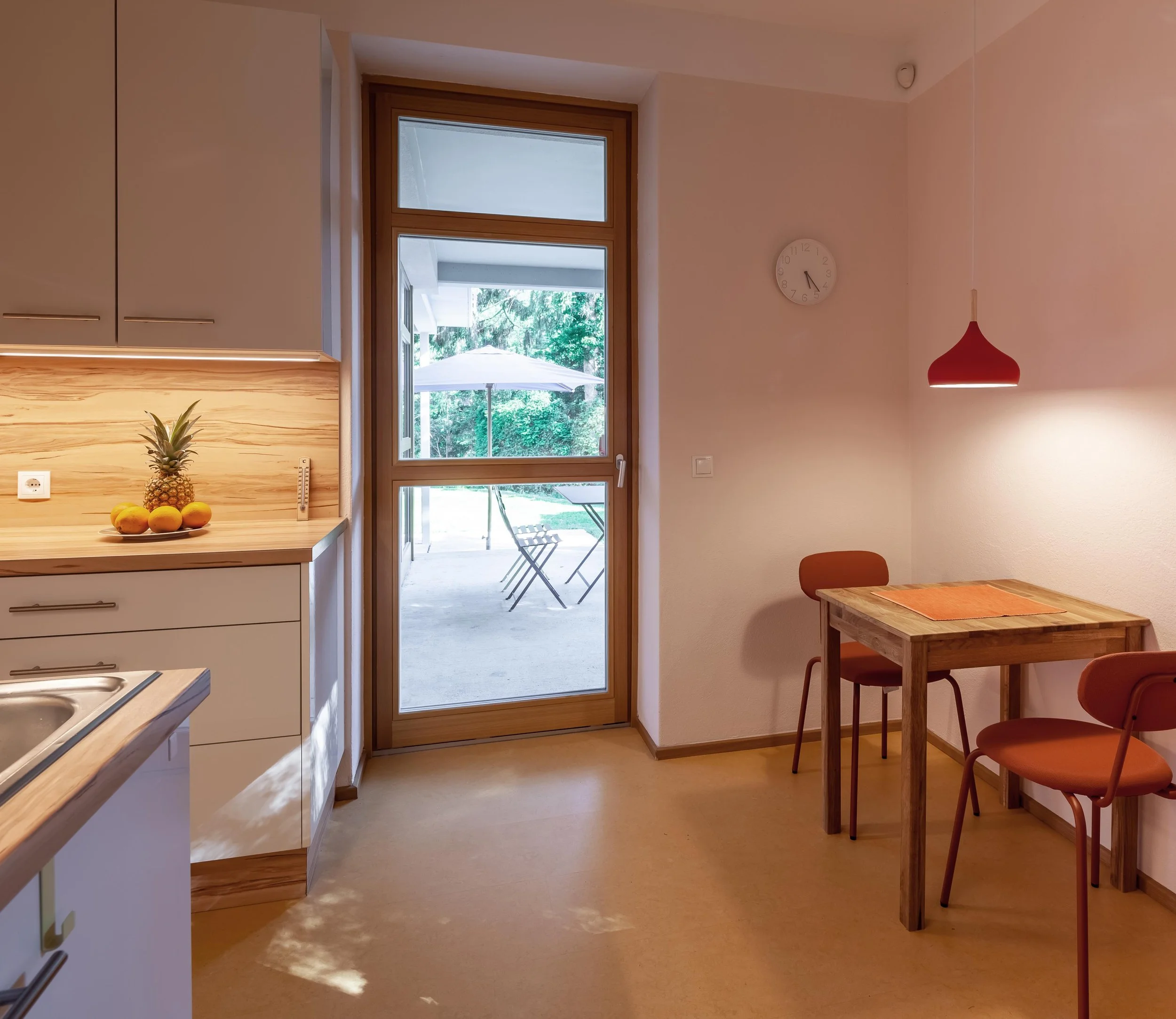 Ein Esszimmer mit Holz-Tisch und roten Stühlen, eine Tür führt auf eine Terrasse mit Sonnenschirm und Garten. Das Zimmer ist schlicht mit weißer Wand, einer roten Pendelleuchte und einer weißen Uhr.