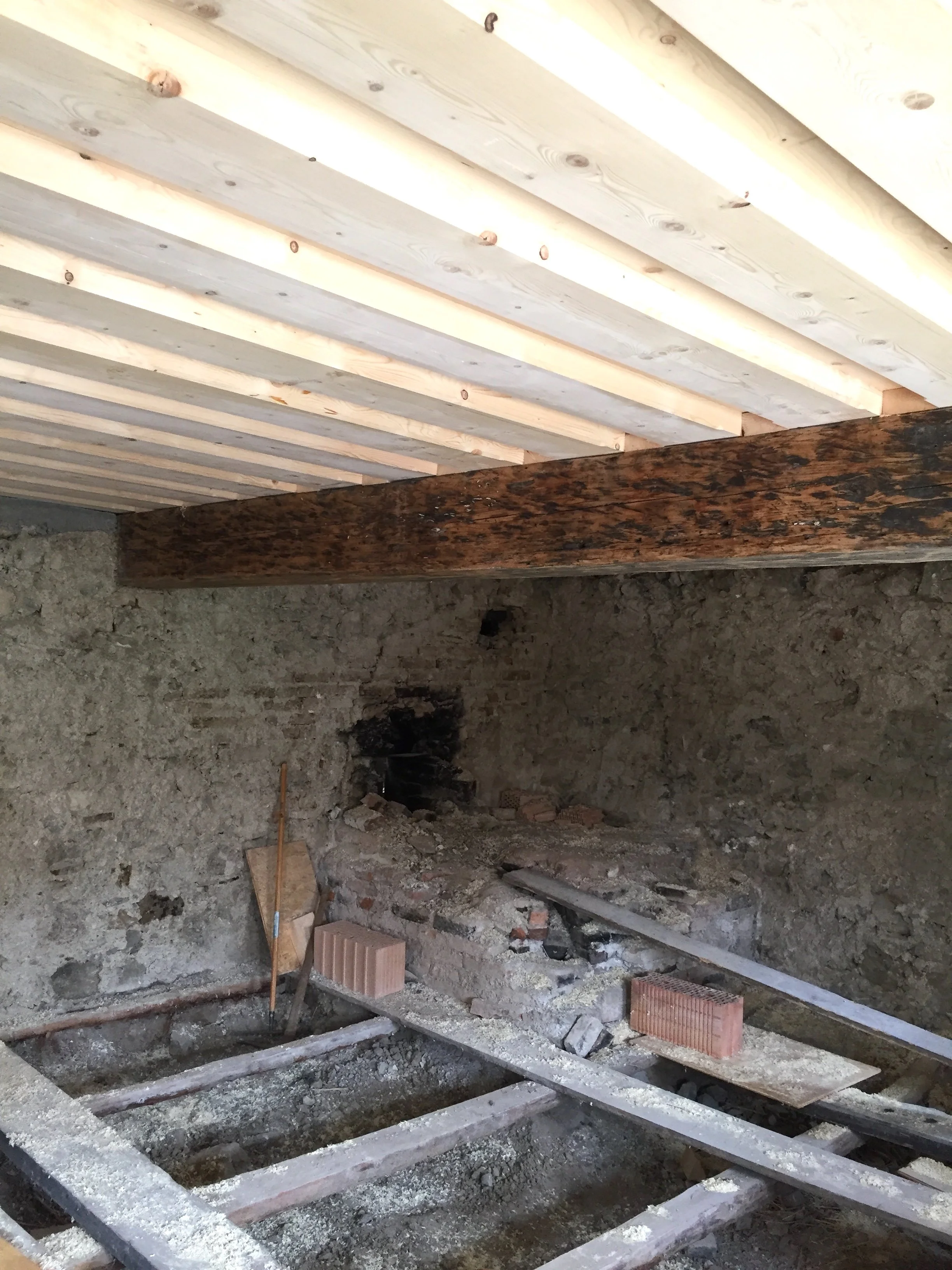 Blick in einen Bau- oder Renovierungsraum mit unvollständigem Boden, eine Holzbalken-Decke und eine Backsteinmauer mit Baustoffen und Werkzeugen.