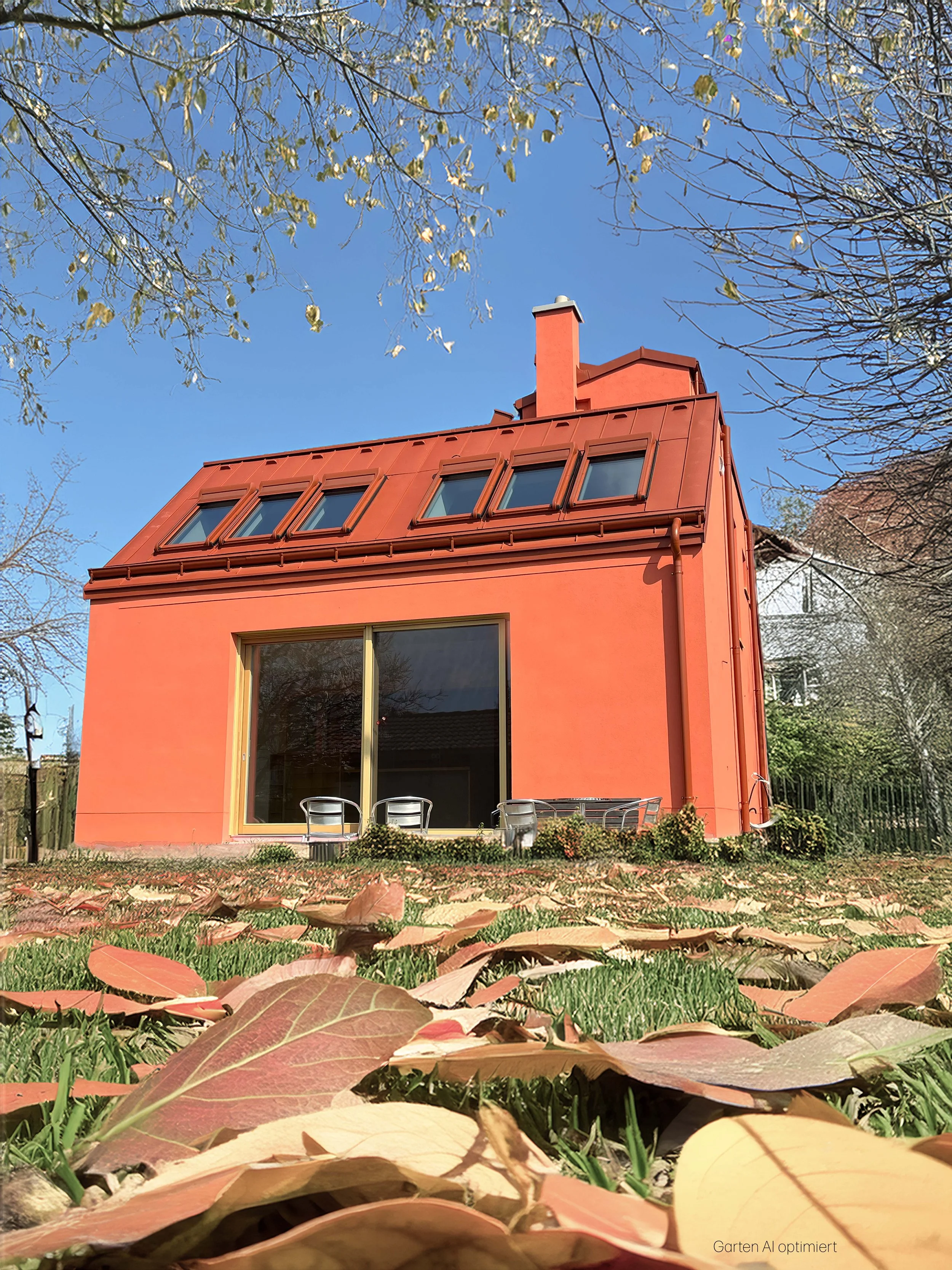 Ein lachsrotes, zweistöckiges Haus mit großen Fenstern und Schornstein, umgeben von Bäumen mit herabfallenden Blättern, unter blauem Himmel.