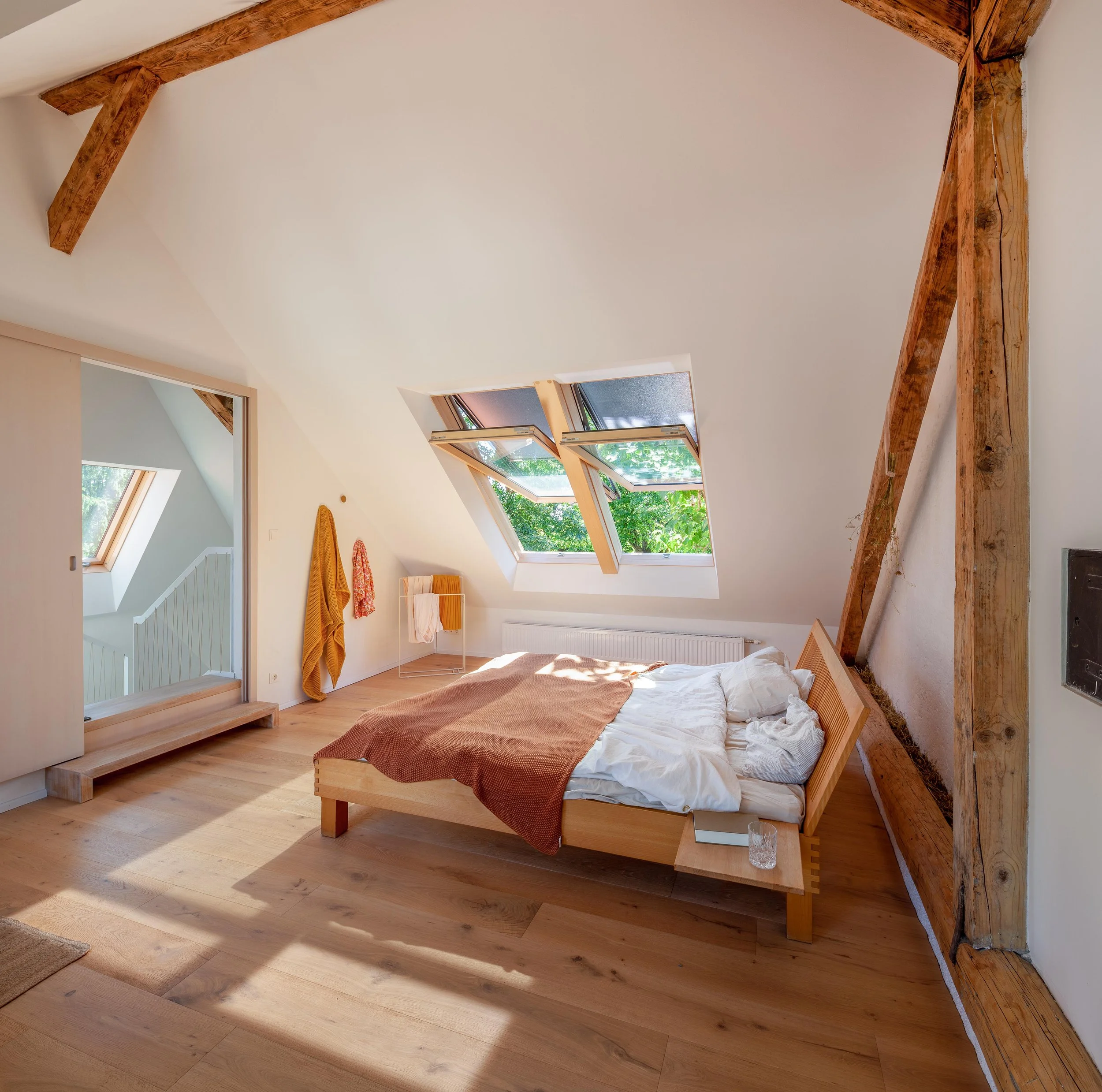 Schlafzimmer mit Holzbettrahmen, großen Dachfenstern, Holzbalken und Lichteinfall, in einem modernen, hellen Raum.