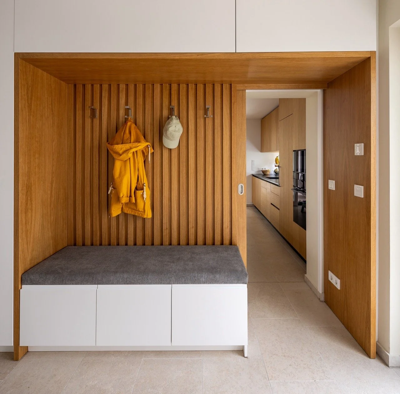 Ein Eingangsbereich mit einer Holz-Wandverkleidung und Haken, an denen eine gelbe Jacke und zwei Hüte hängen. Davor befindet sich eine Sitzbank mit grauem Kissen. Im Hintergrund ist eine offene Küche mit Holzmöbeln zu sehen.