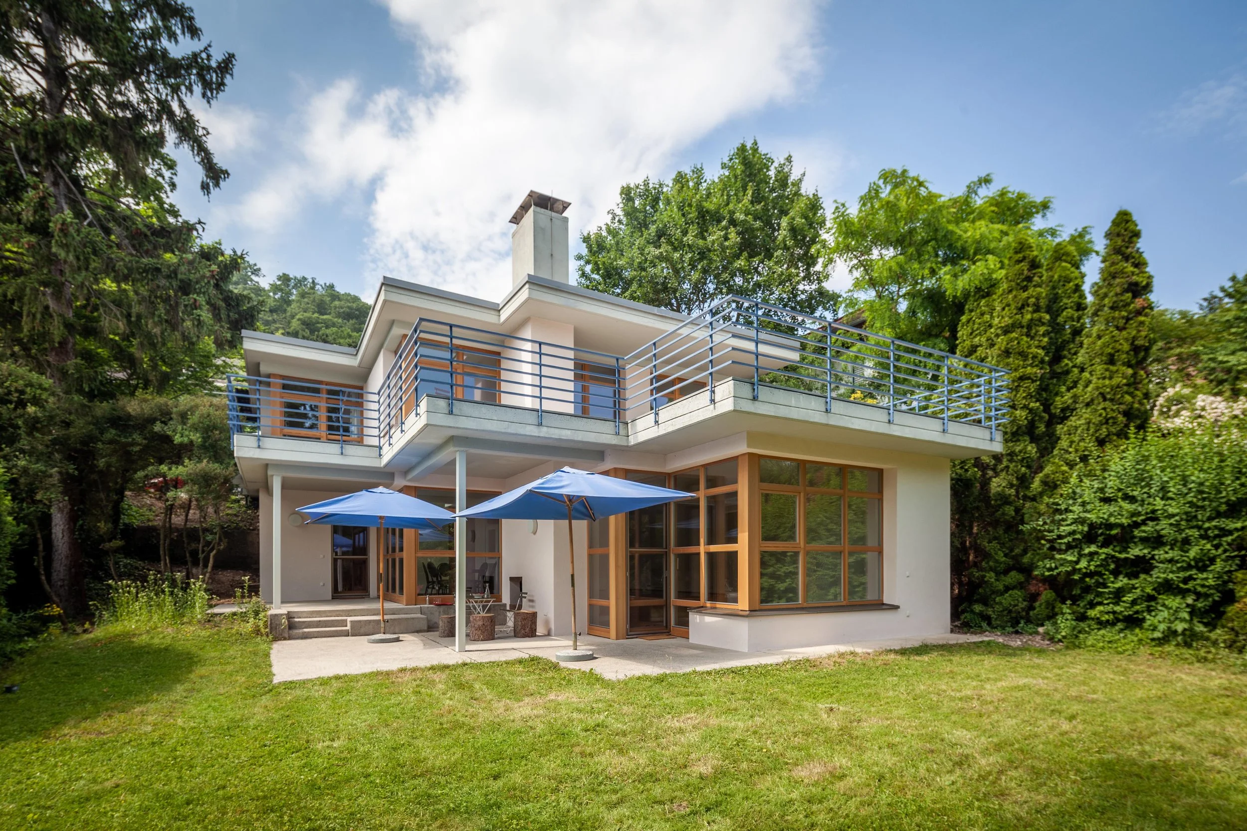 Modernes Haus mit großen Fenstern, Balkon und Garten, Sonnenschirme im Garten, umgeben von Bäumen, blauer Himmel.