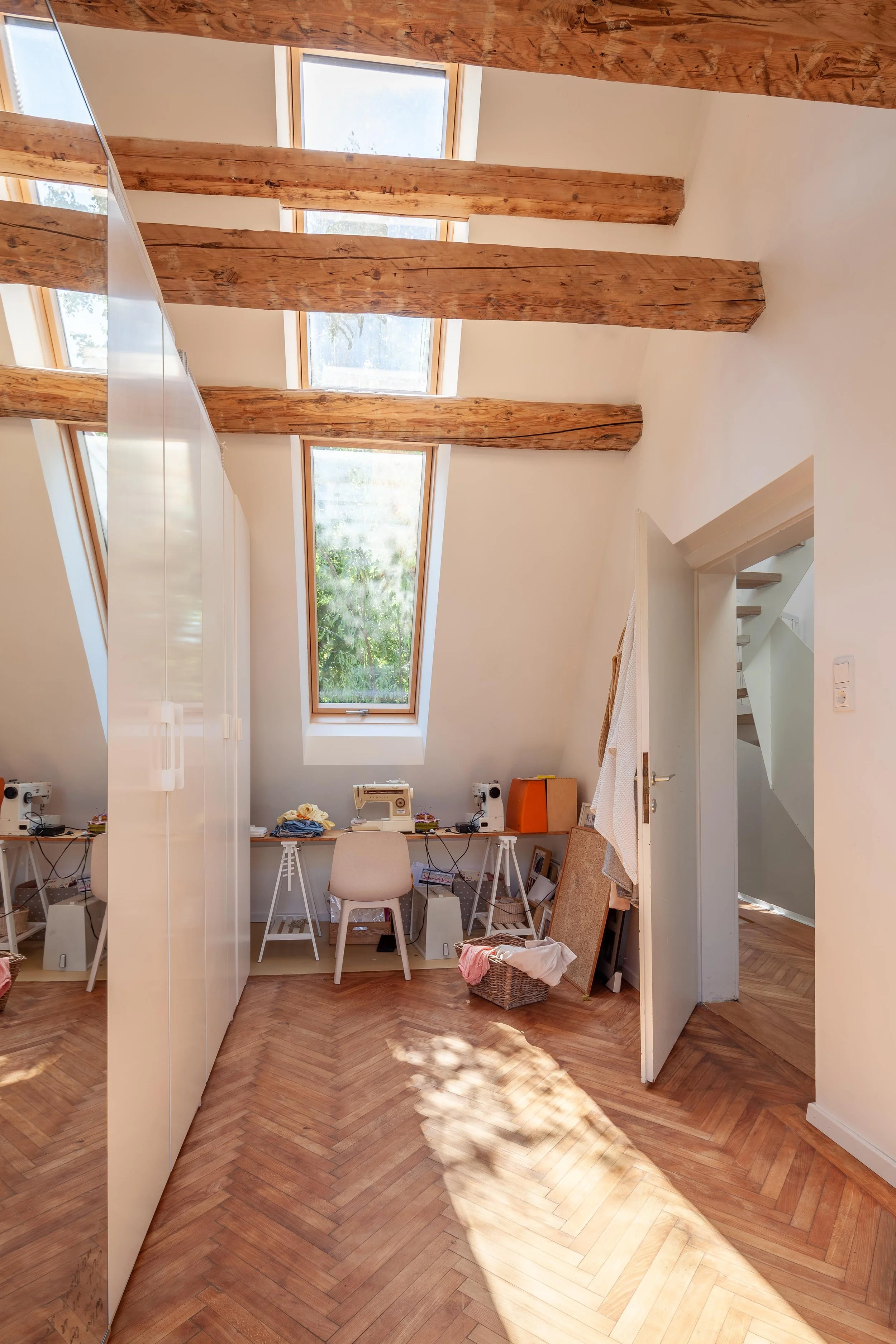 Ein heller Raum mit Holzdielenboden, schrägen Deckenbalken und mehreren Dachfenstern. Es gibt eine weiße Schrankfront, einen Schreibtisch mit Nähmaschinen, Kleidungsstücken und verschiedenen Gegenständen.