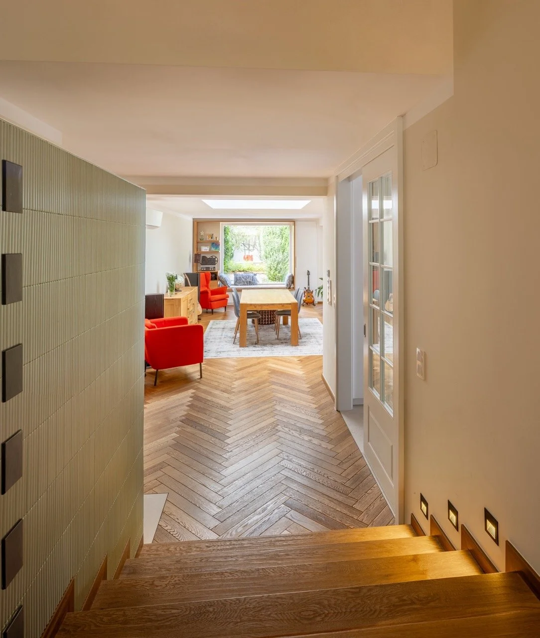 Einen Flurbereich, blickt in einen offenen Wohn- und Essbereich mit Holzboden, roten Sesseln, einem Esstisch, großen Fenster mit Blick auf einen Garten, und Holzboden im Hintergrund.