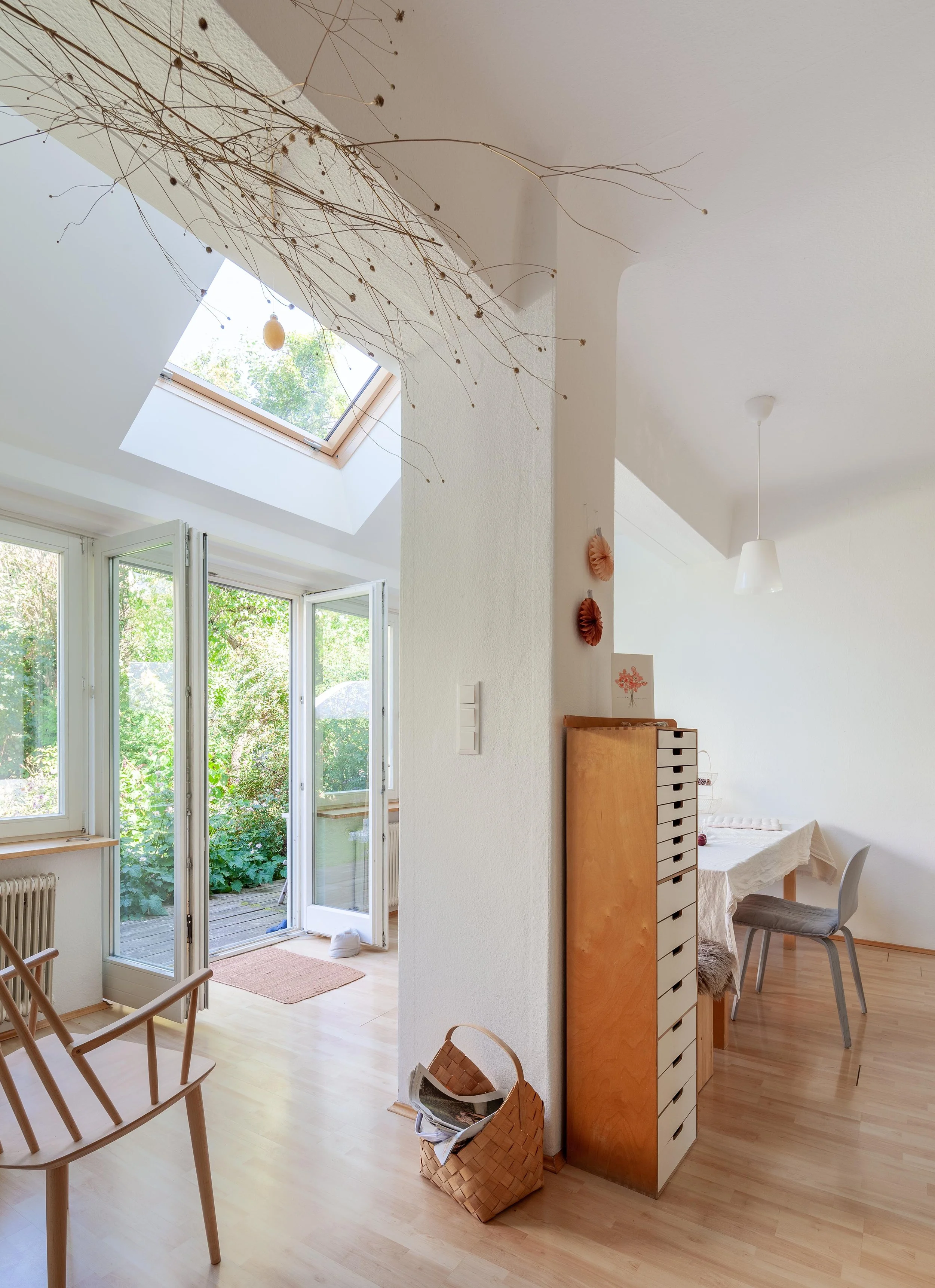 Ein helles Wohnzimmer mit offenen französischen Türen, die auf eine grüne Terrasse führen. Ein Dachfenster lässt Tageslicht herein. Es gibt einen kleinen Tisch mit Stühlen, einen hohen Holzschrank mit mehreren Schubladen, eine Papierdekoration an der