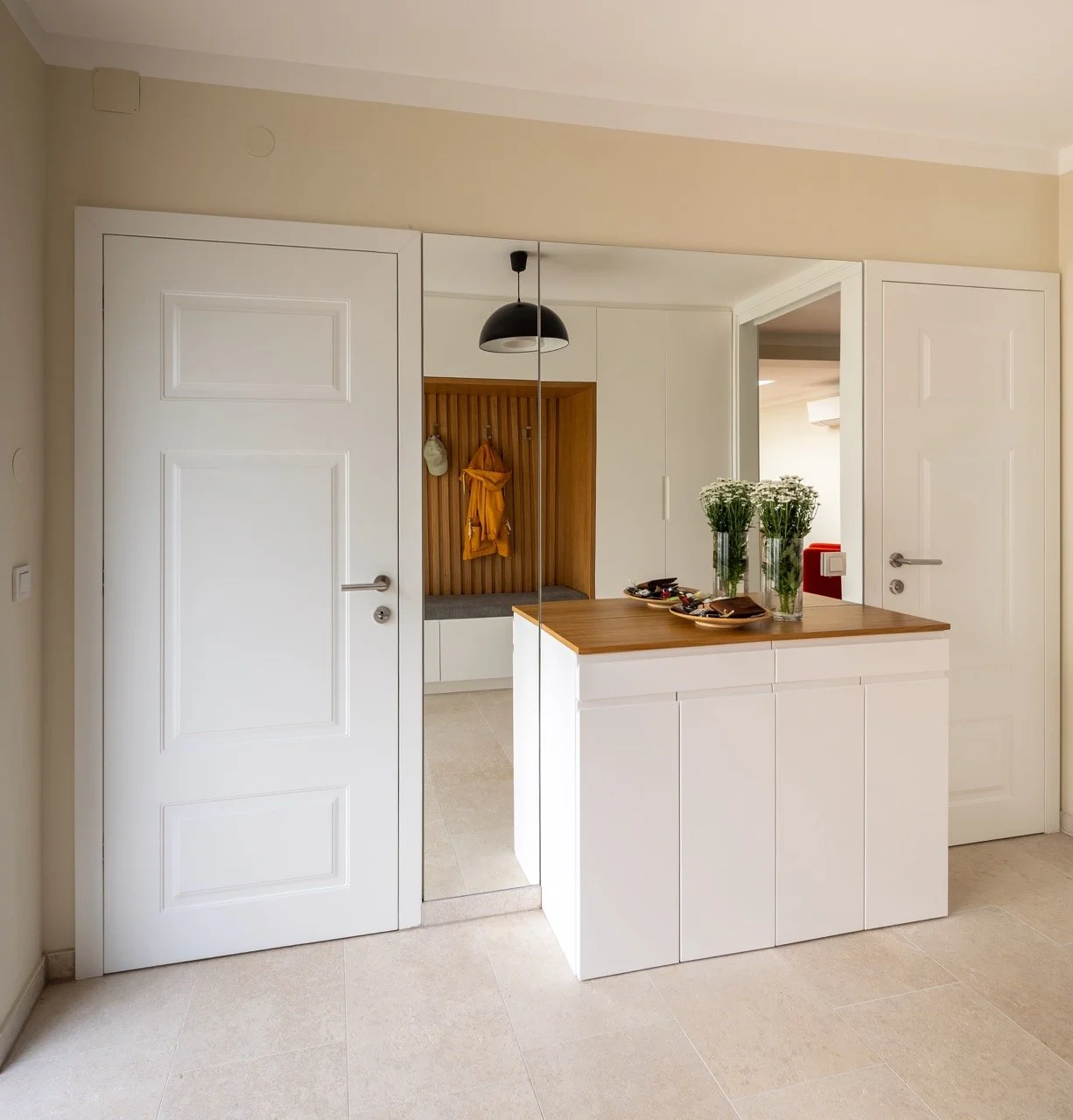 Ein moderner Eingangsbereich mit weißen Türen, einer Holzarbeitsplatte, Vasen mit weißen Blumen und einem Spiegel, der die Szene reflektiert.