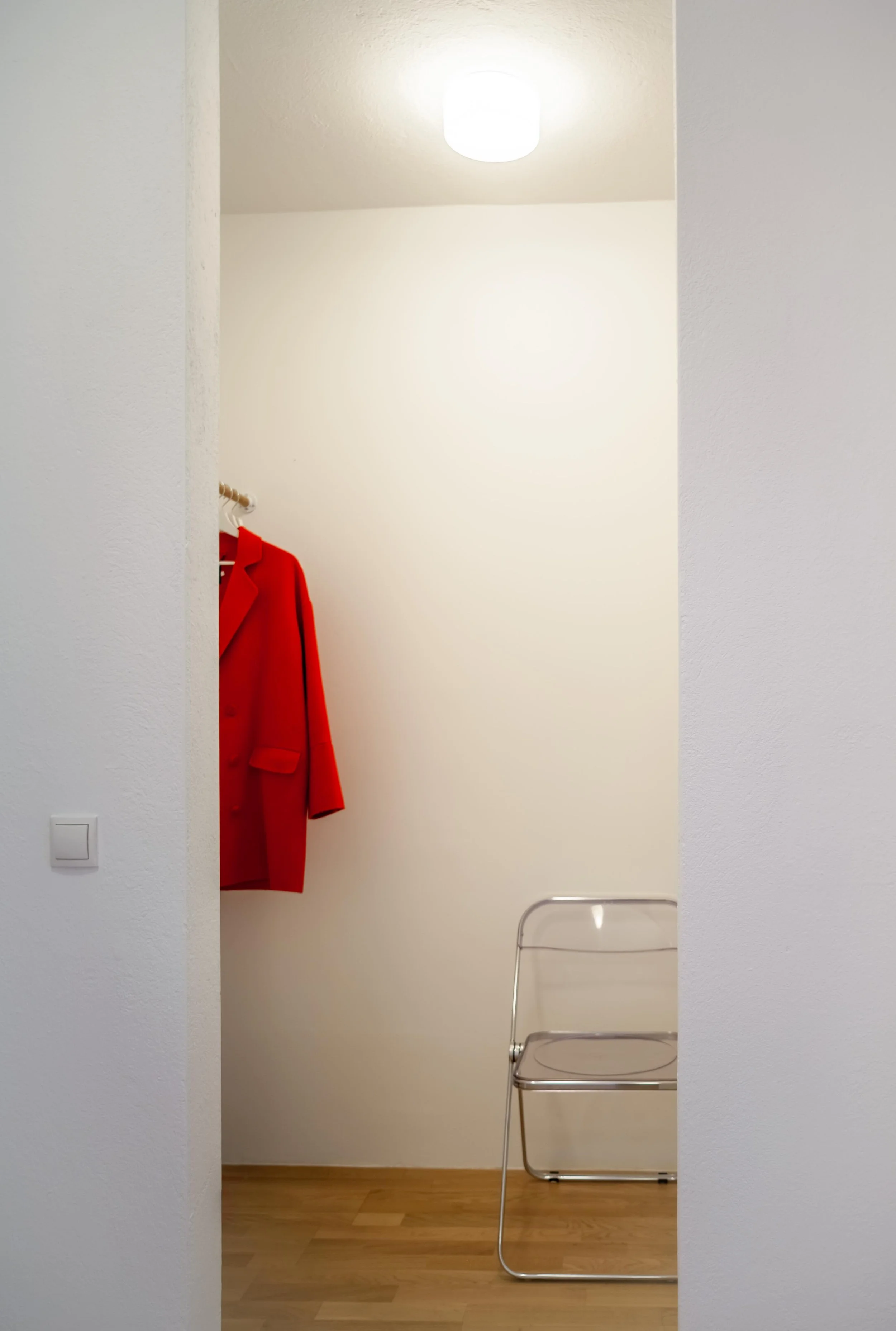 Ein heller, minimalistisch eingerichteter Raum mit weißer Wand, einer roten Jacke an einem Garderobenhaken und einem silbernen Stuhl auf Holzfußboden.