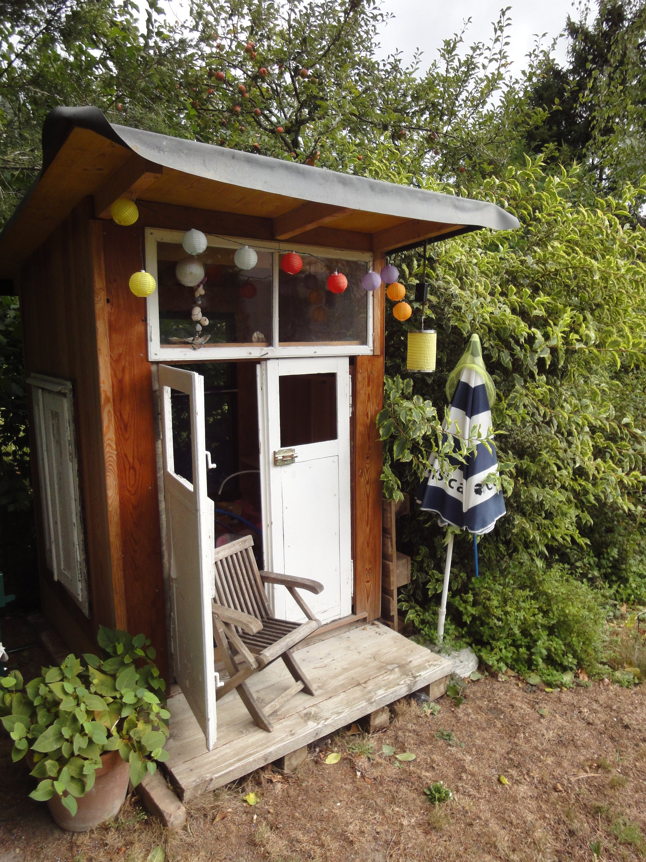 Kleine Holzhütte im Garten mit bunten Laternen und Sonnenschirm, umgeben von Büschen und Bäumen.