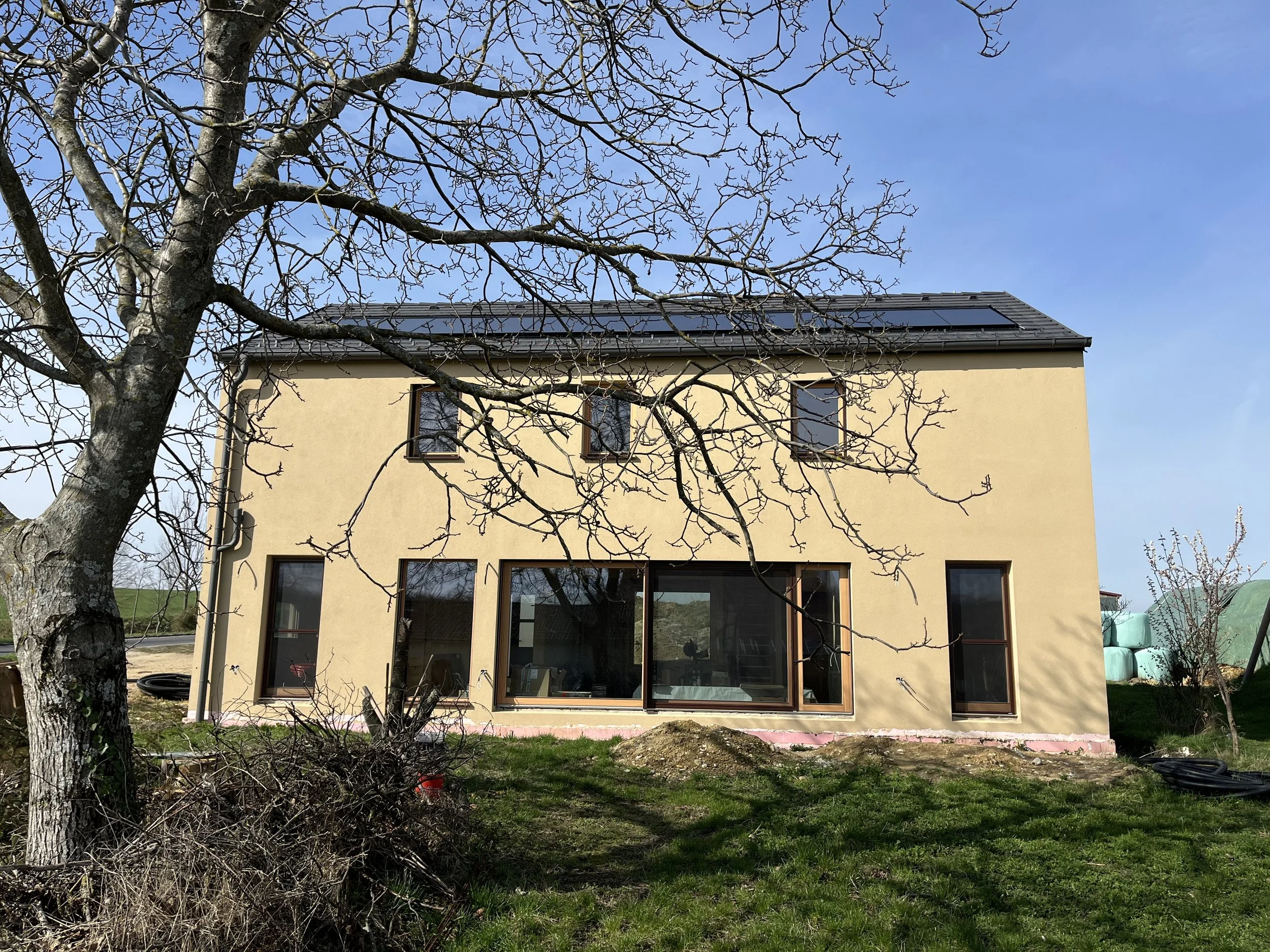 Neues Beiges Haus mit großen Fenstern, im Frühling ohne Blätter an den Bäumen davor, auf einer grünen Rasenfläche, blauer Himmel.