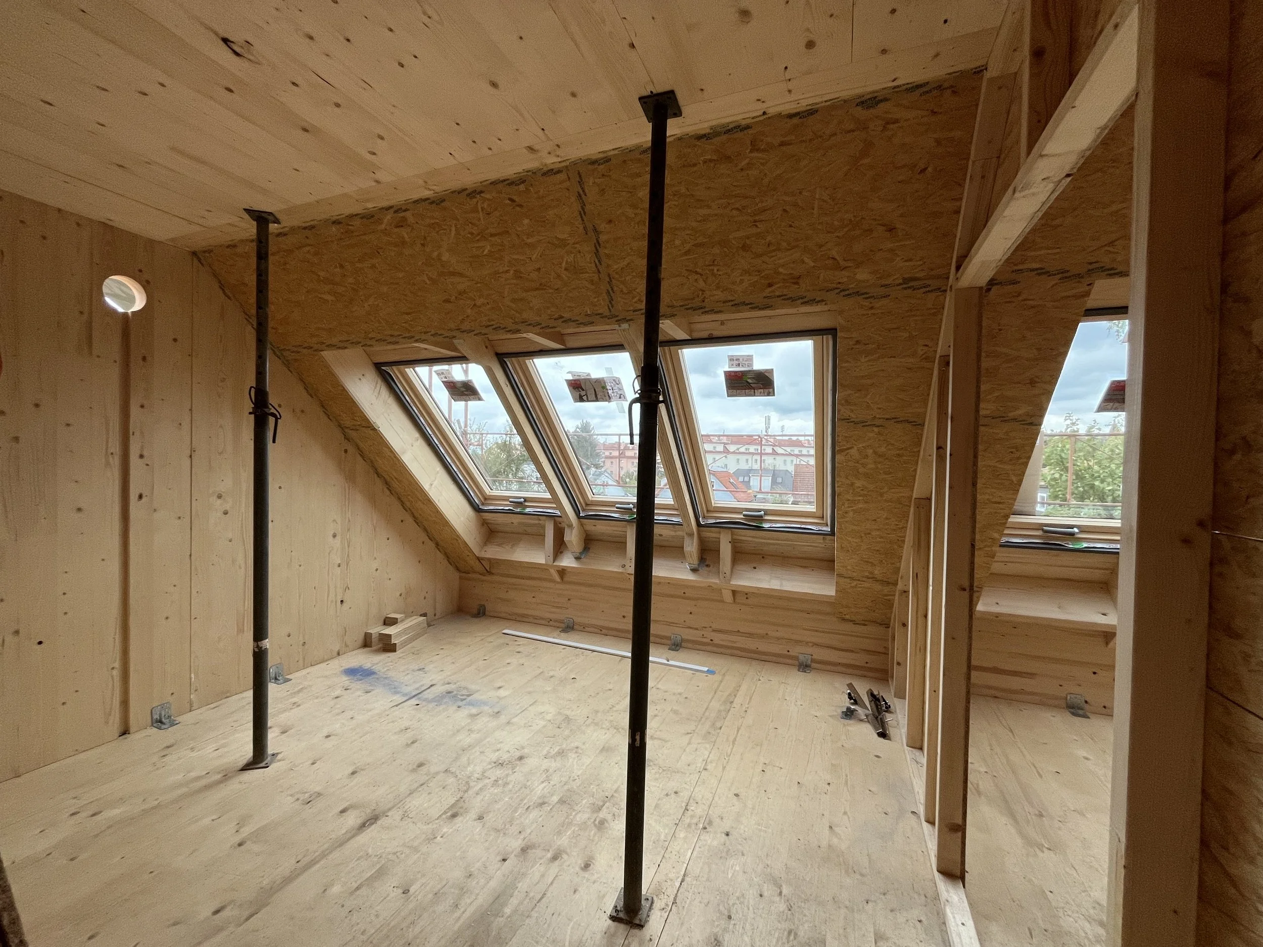 Innenraum eines Dachgeschosses im Bau, mit Dachfenstern und rohem Holzbau. Temporäre Stützstangen sind sichtbar.