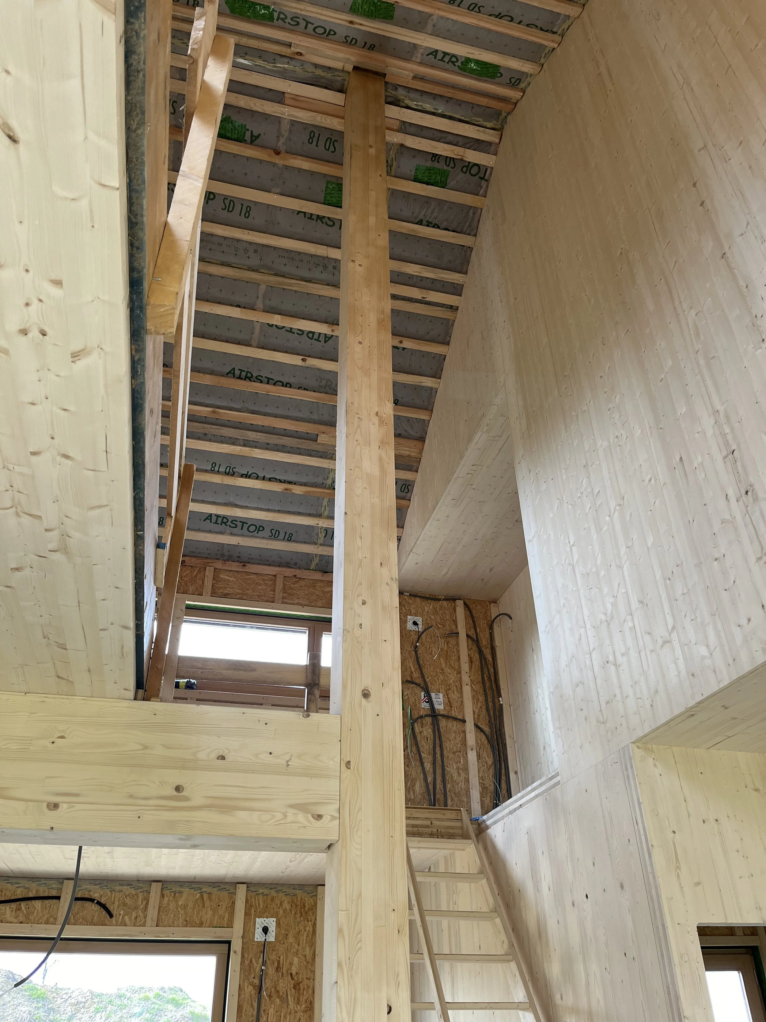 Innenansicht eines Holzhauses im Bau, mit sichtbarer Dachkonstruktion, Massivholzwänden, Leitungen und einer Leiter.