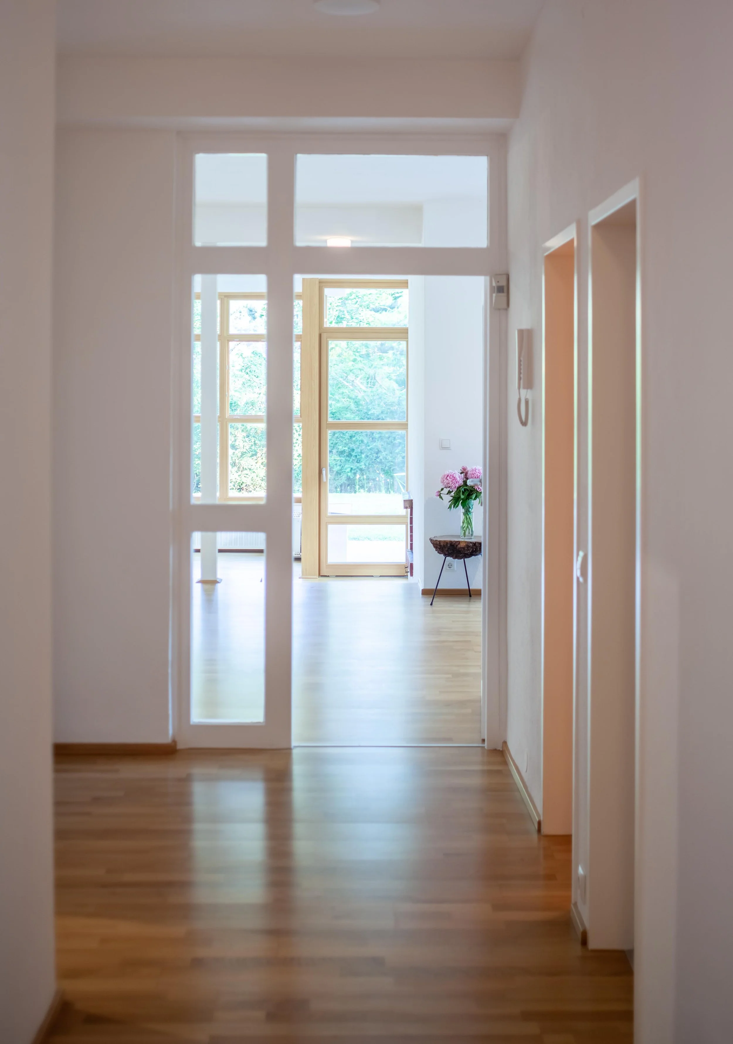Blick in einen hellen Flur mit Holzboden, durch eine Glastür ist ein Wohnzimmer mit großen Fenstern und einer Tür nach draußen sichtbar, im Raum steht ein Tisch mit einem Blumenstrauß.