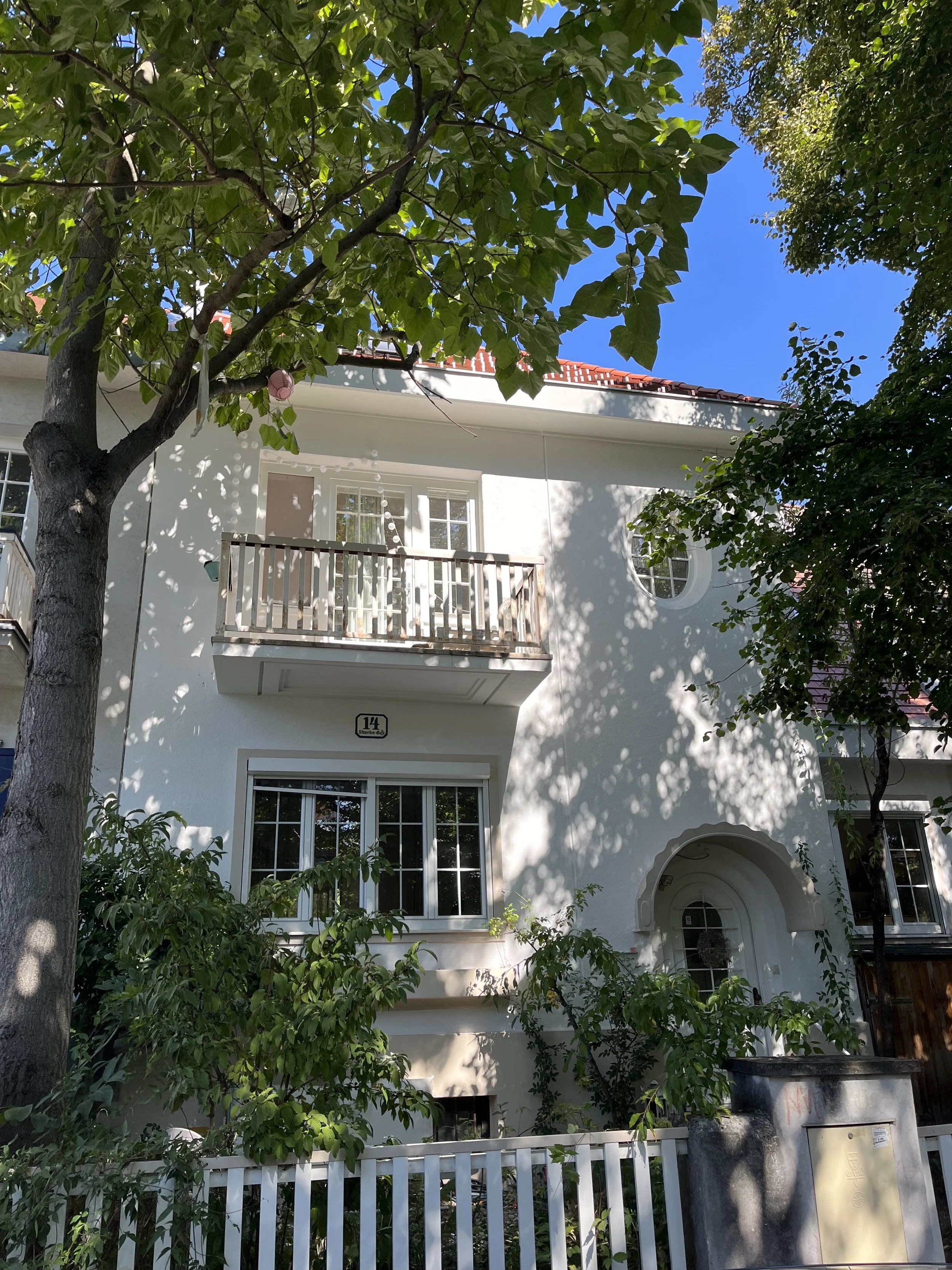 Weißes zweistöckiges Haus mit Balkon, grünen Büschen und Baum, blaulastiger Himmel, Schatten der Blätter auf der Fassade, mit Gitterzaun im Vordergrund.