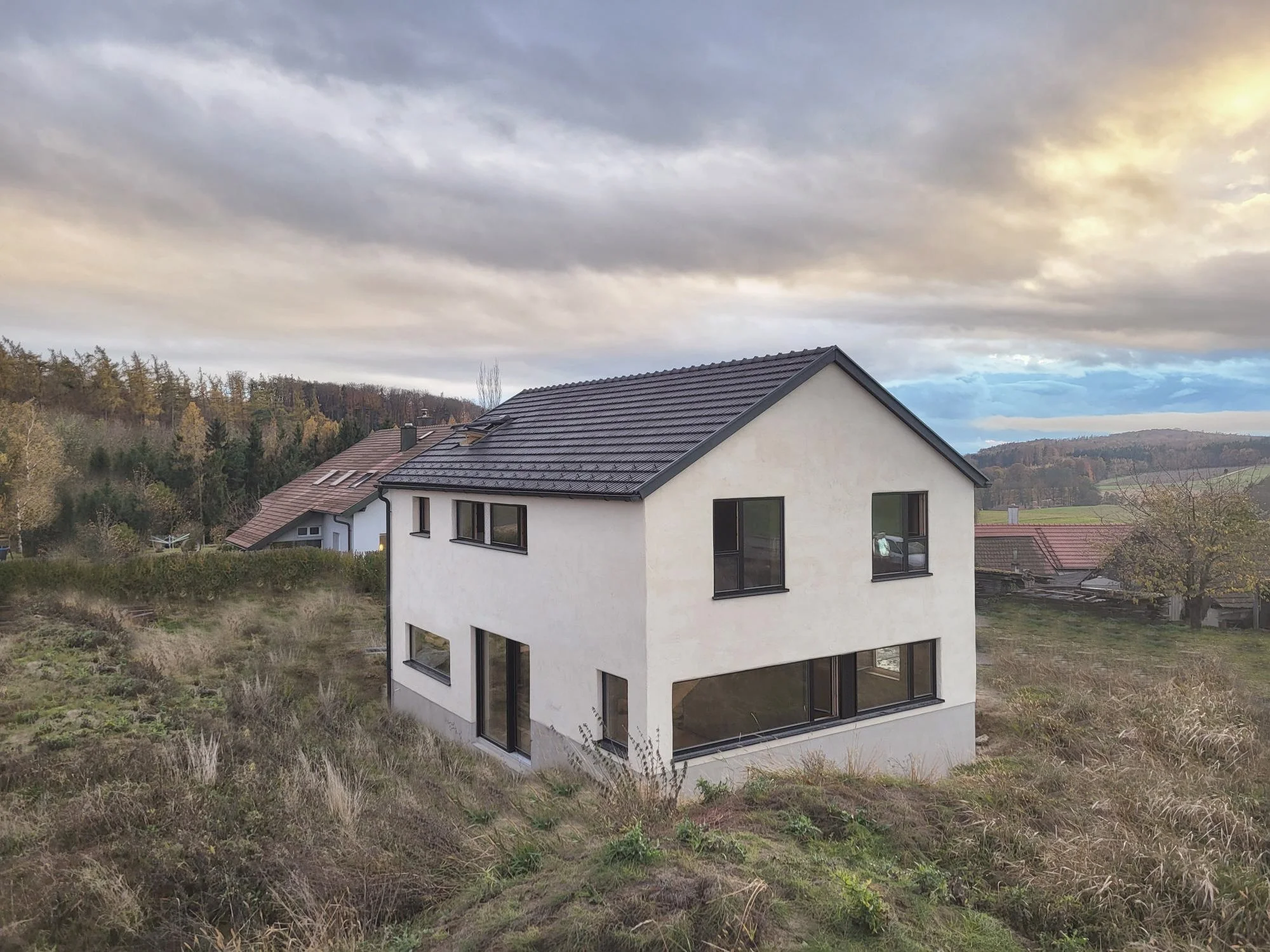 Modernes zweistöckiges weißes Haus mit Kalkputzfassade in ländlicher Gegend, umgeben von Hügeln und Wäldern, mit mehreren großen Fenstern und dunklem Dach, bei bewölktem Himmel.