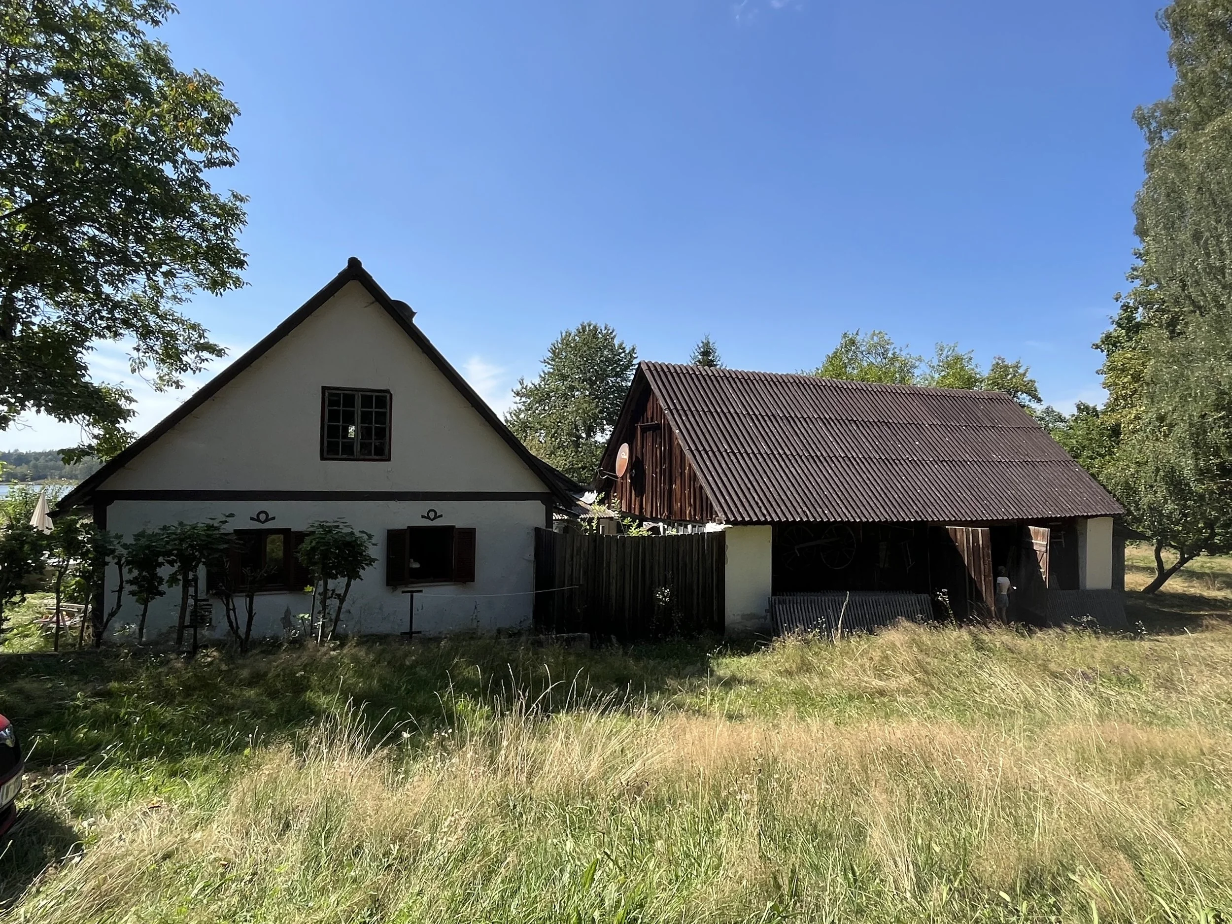 Zwei Bauernhäuser, eines weiß mit dunklen Fensterläden, das andere aus Holz mit rotem Wellblech-Dach, auf einer Wiese unter Bäumen bei sonnigem Himmel.