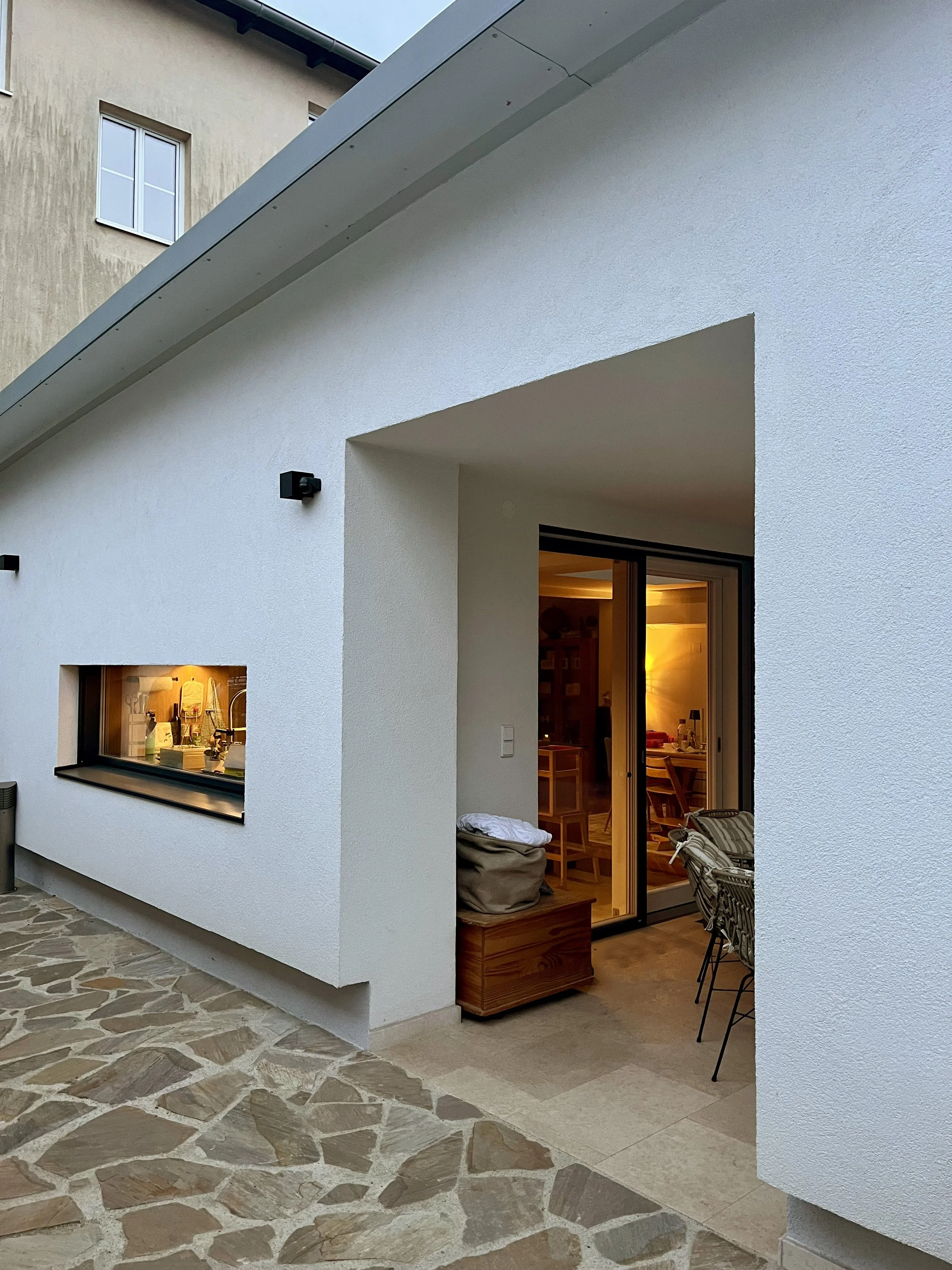 Außenaufnahme eines modernen, weißen Hauses mit offener Terrasse, einblick in einen Wohnraum mit warmem Licht, ein kleiner Fensterraum, Steinboden und Möbel im Innenbereich.