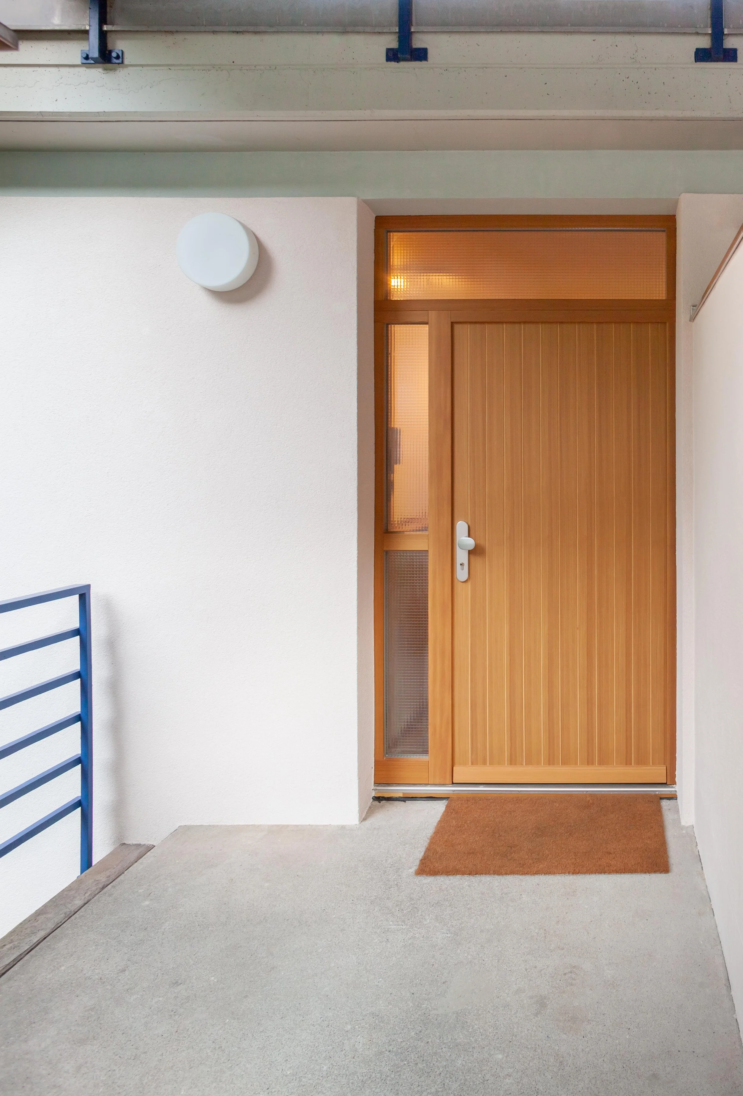 Holztür in einem Eingangsbereich, weiße Wände und eine Außenbeleuchtung.