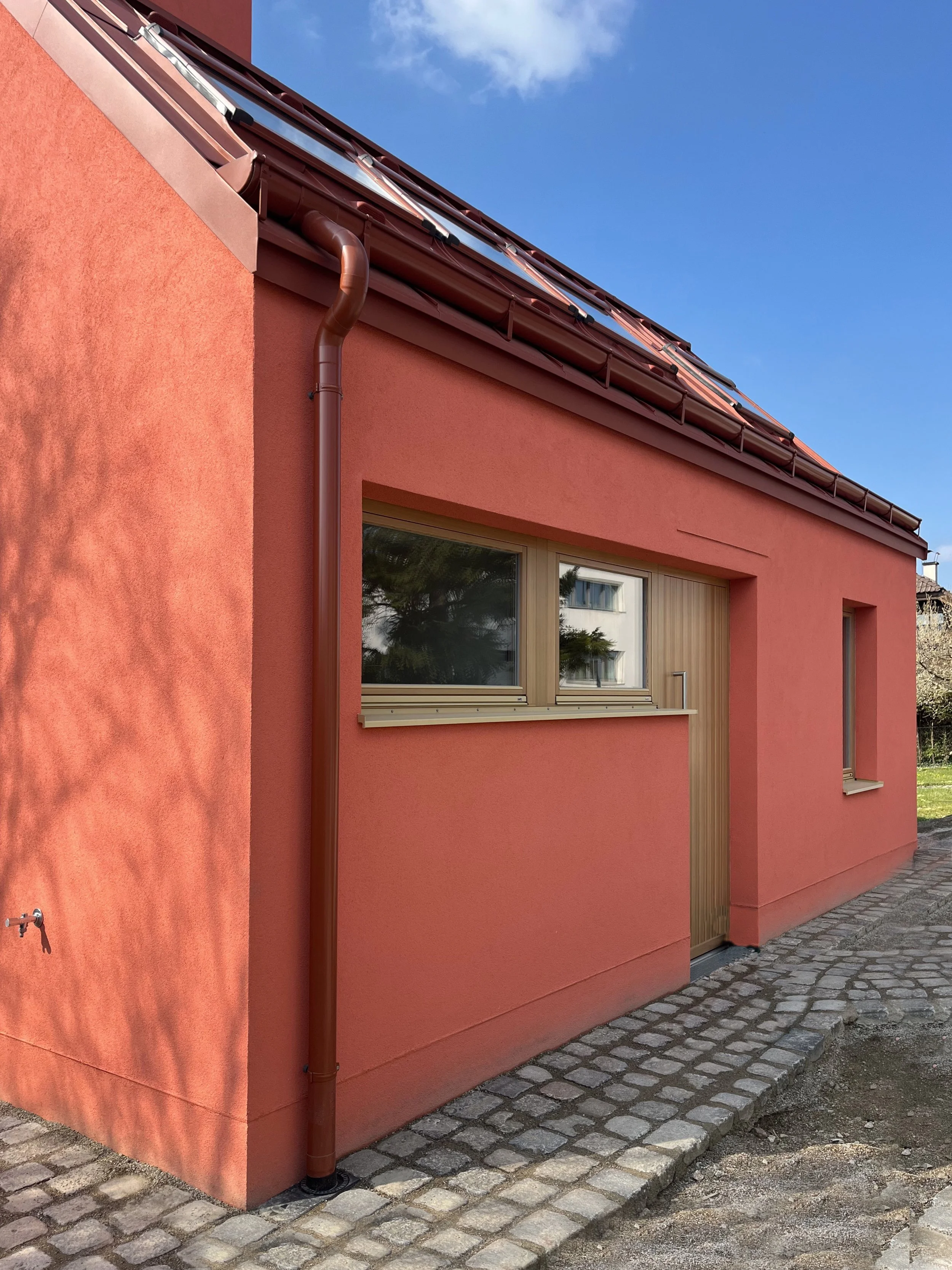 Ein lachsrotes Haus mit rotem Blechdach, zwei Holzfenster, einer Holzeingangstür und einem gepflastertem Weg im Garten, bei sonnigem Himmel.