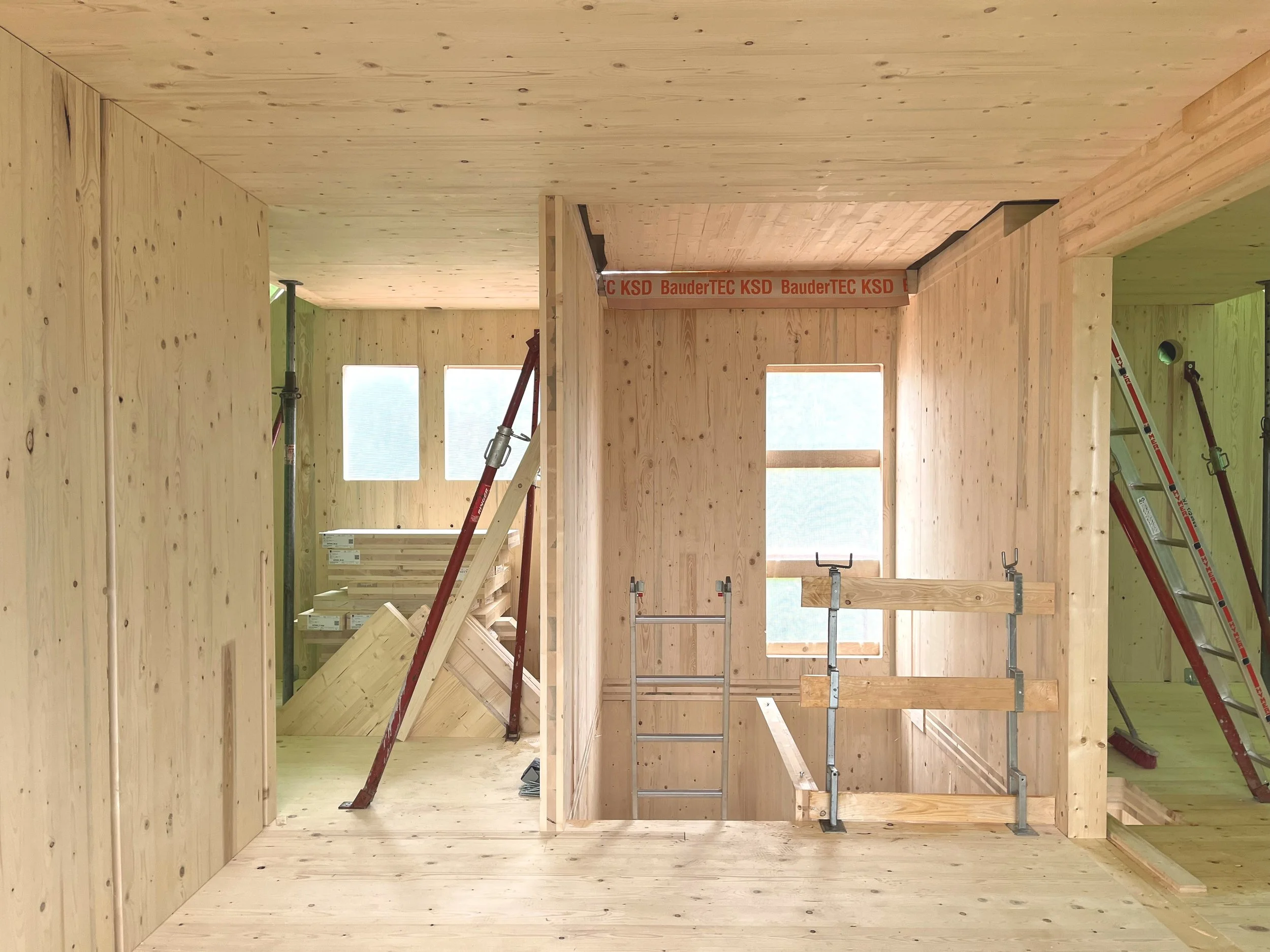 Innenraum eines Holzhauses im Bau, mit Wänden, Decken und Treppenkonstruktion aus Holz, sowie Leitern und Baumaschinen.