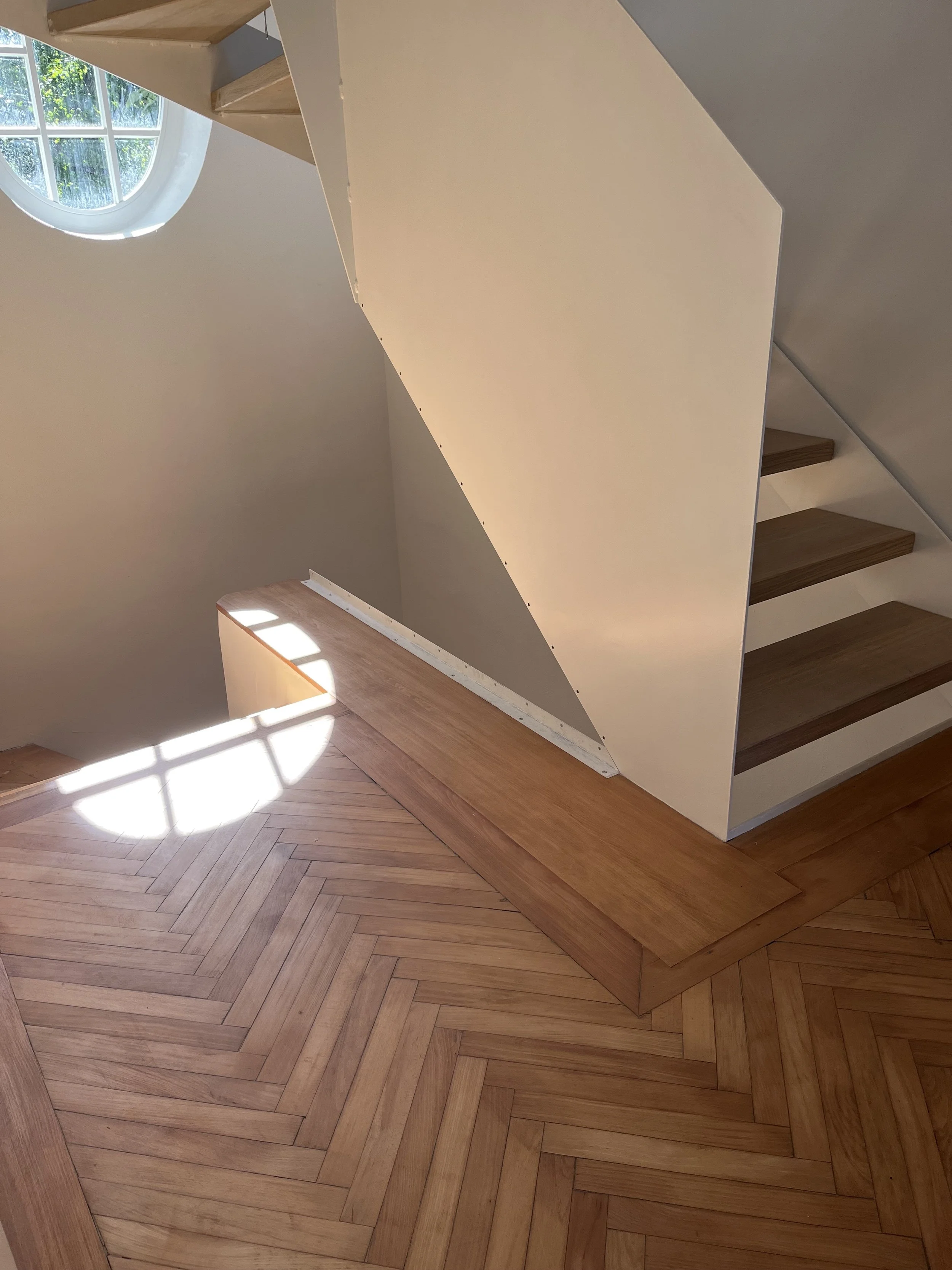 Innenaufnahme eines Treppenhauses mit einem runden Fenster, Holzdielenboden im Fischgrätmuster und einer halb offenen Treppe mit Holzstufen und weißen Wänden.