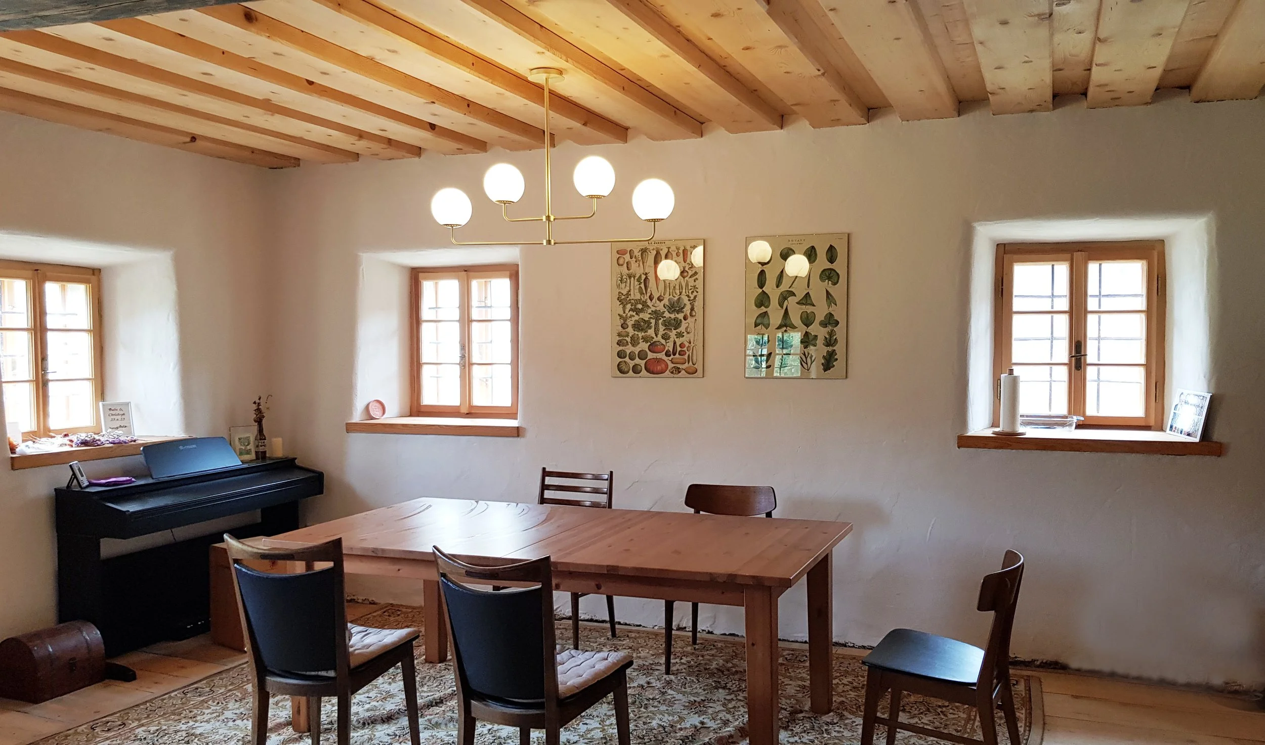 Gemütlicher Esszimmerraum mit Holztisch, sechs Stühlen, zwei Fenstern, einer Deckenlampe und einem Keyboard in einer rustikalen, hell gestrichenen Umgebung mit Lehmputz und Kalkfarbe.