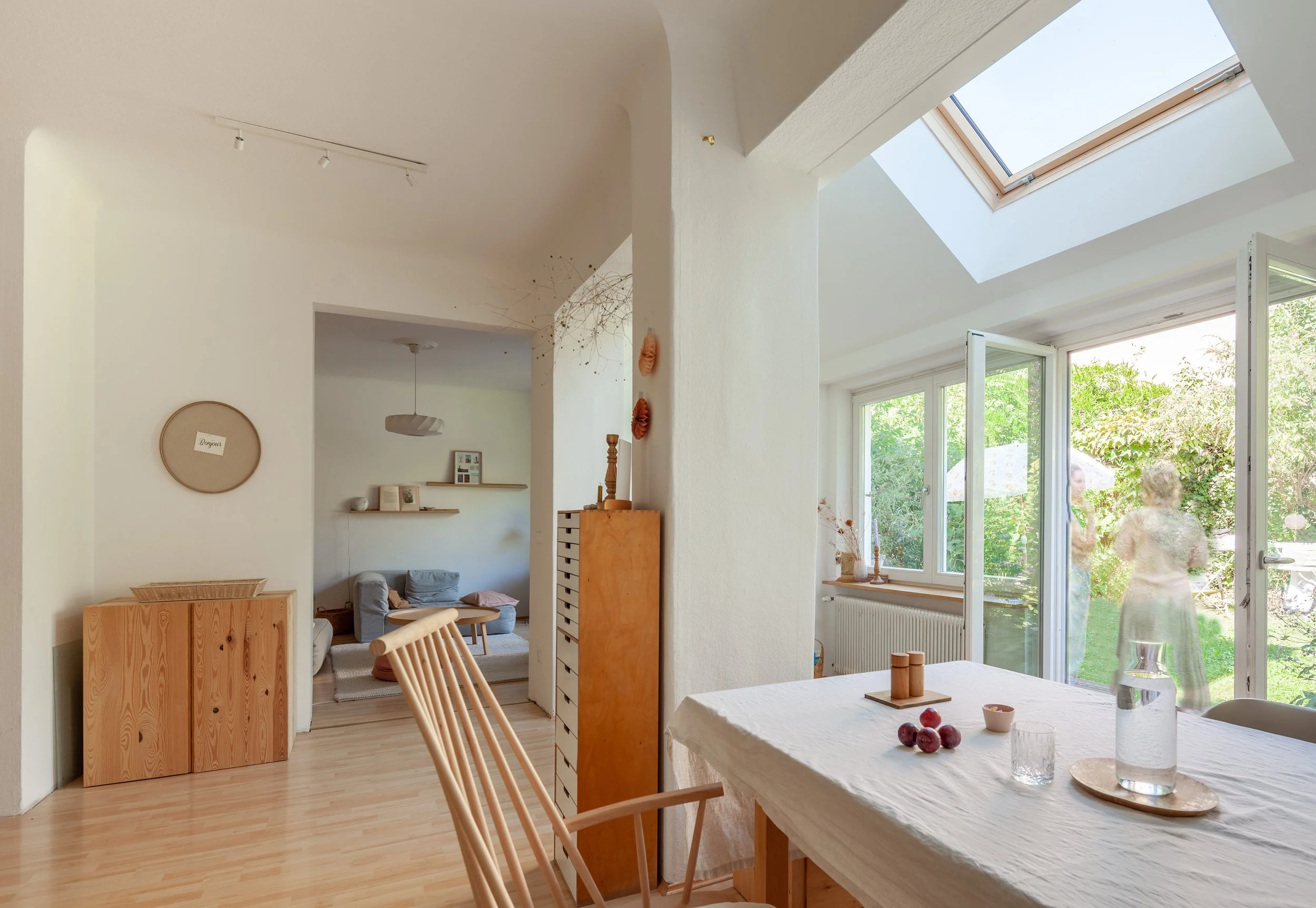 Ein helles Wohnzimmer mit großen Fenstern und einem Blick auf einen Garten. Es gibt einen Esstisch mit einem weißen Tuch, auf dem eine Wasserkaraffe, Gläser und Früchte stehen. Im Hintergrund ist eine Frau im Garten sichtbar.