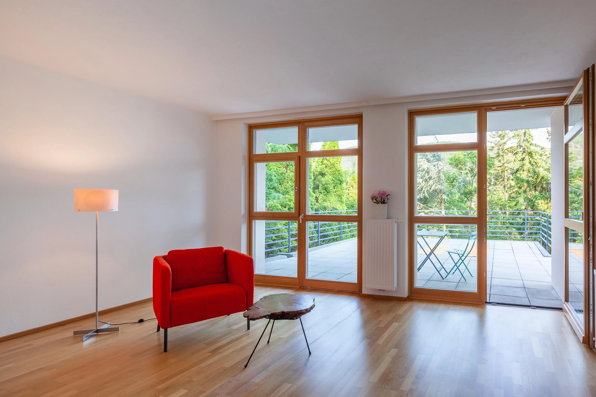 Gemütliches Wohnzimmer mit großer Fensterfront, rotem Sofa, Stehlampe und Balkon mit Blick auf grüne Bäume