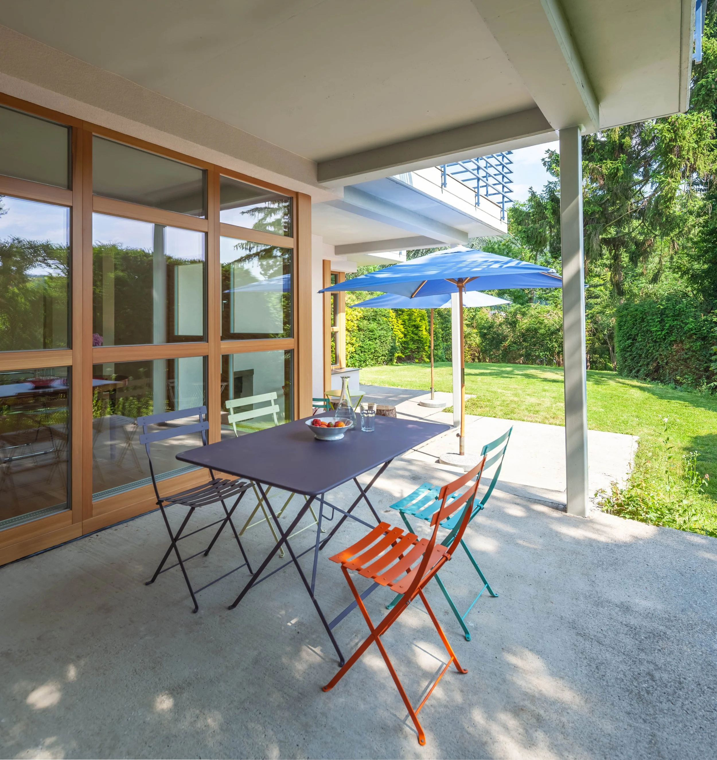 Ein Balkon mit bunten Stühlen, einem Tisch mit Obst und zwei Sonnenschirmen, umgeben von Grünflächen.
