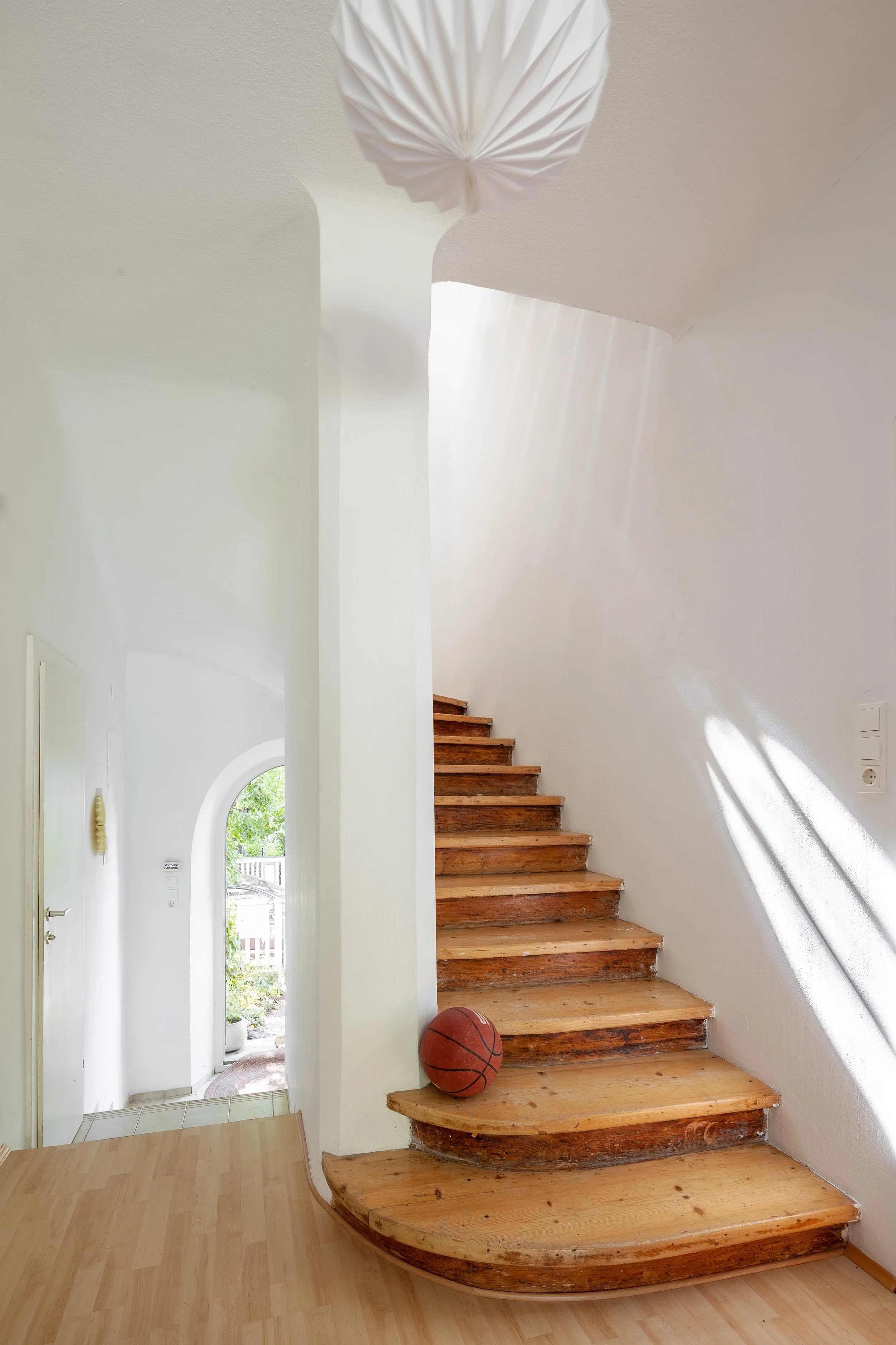 Innenaufnahme eines Hauses mit Treppe, ein Basketball auf der Treppe, weiße Wände, Beleuchtung, durch die Tür blickt man nach draußen auf einen Garten mit Bäumen.