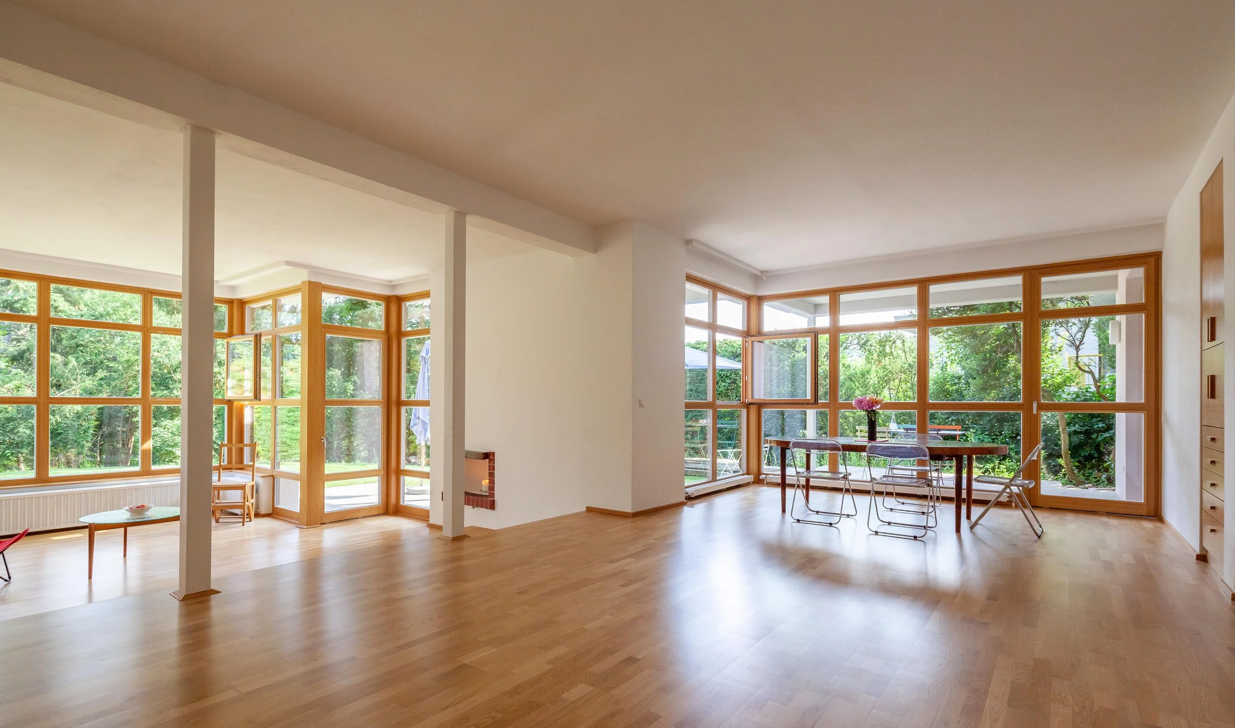 Helles, offenes Wohnzimmer mit großen Fenstern und Holzboden, Esstisch mit Stühlen, im Hintergrund grüne Natur sichtbar.