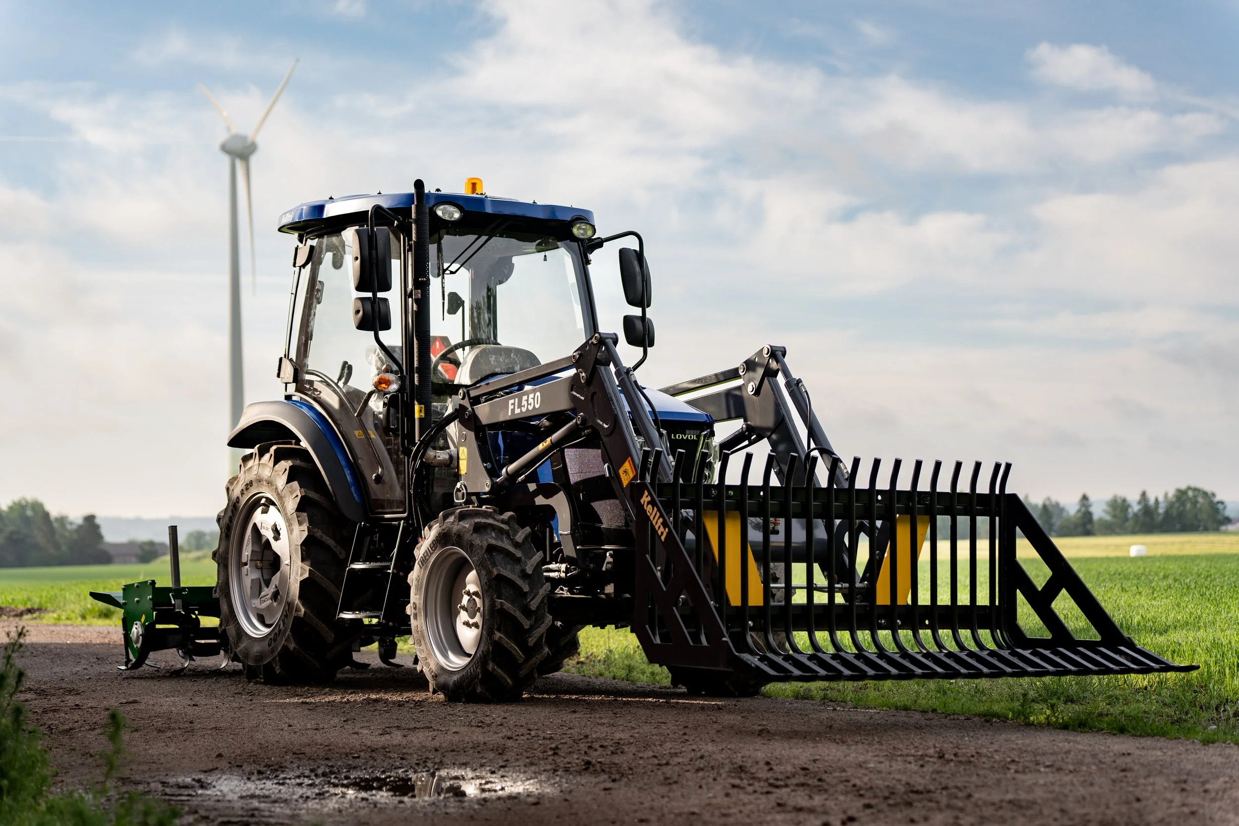 A tractor with a front loader attachment on a dirt path in a field with green grass, with a wind turbine and cloudy sky in the background.