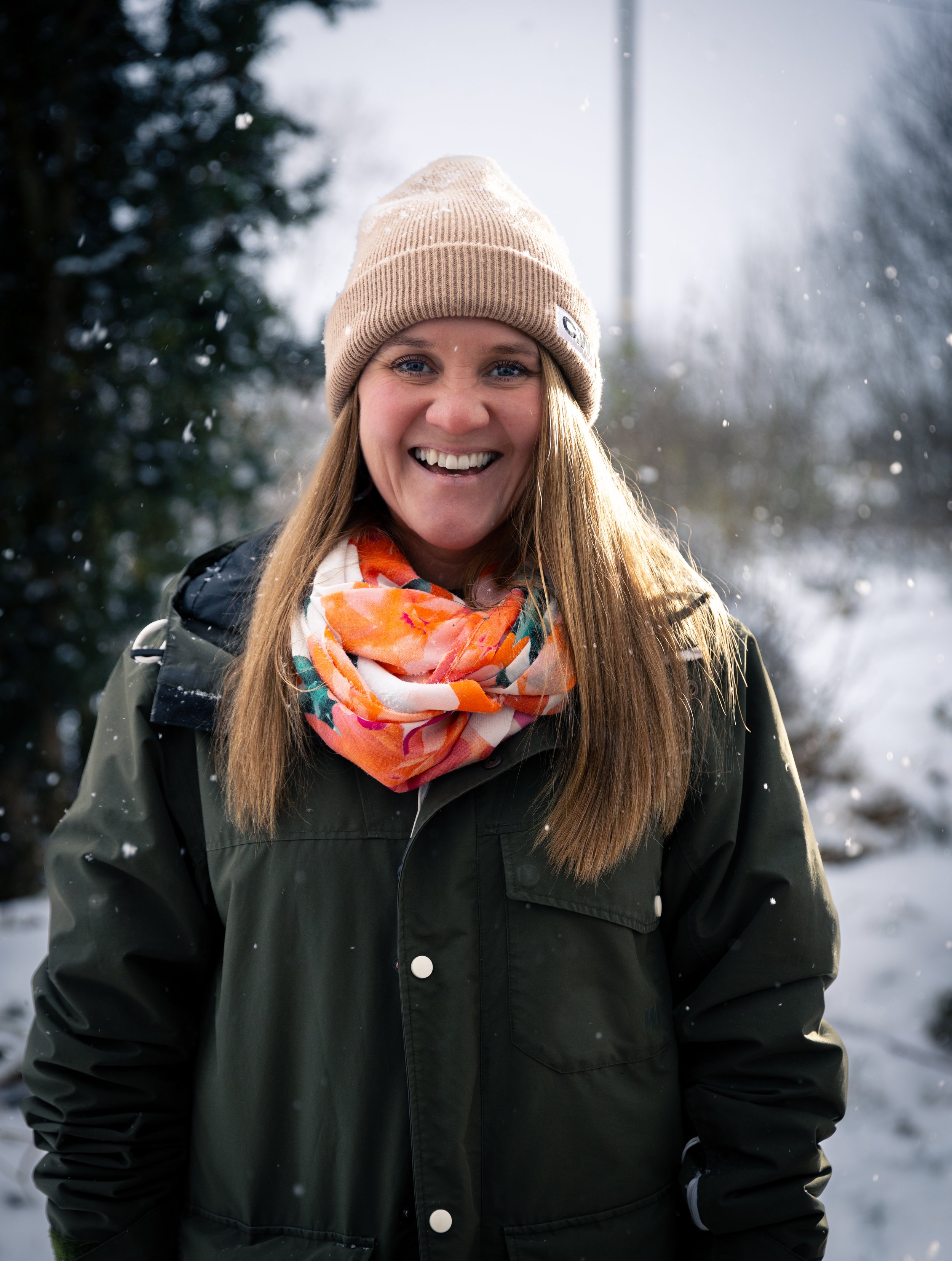 A smiling woman with long hair wearing a beige beanie, a black jacket, and a colorful scarf outdoors in a snowy winter landscape.