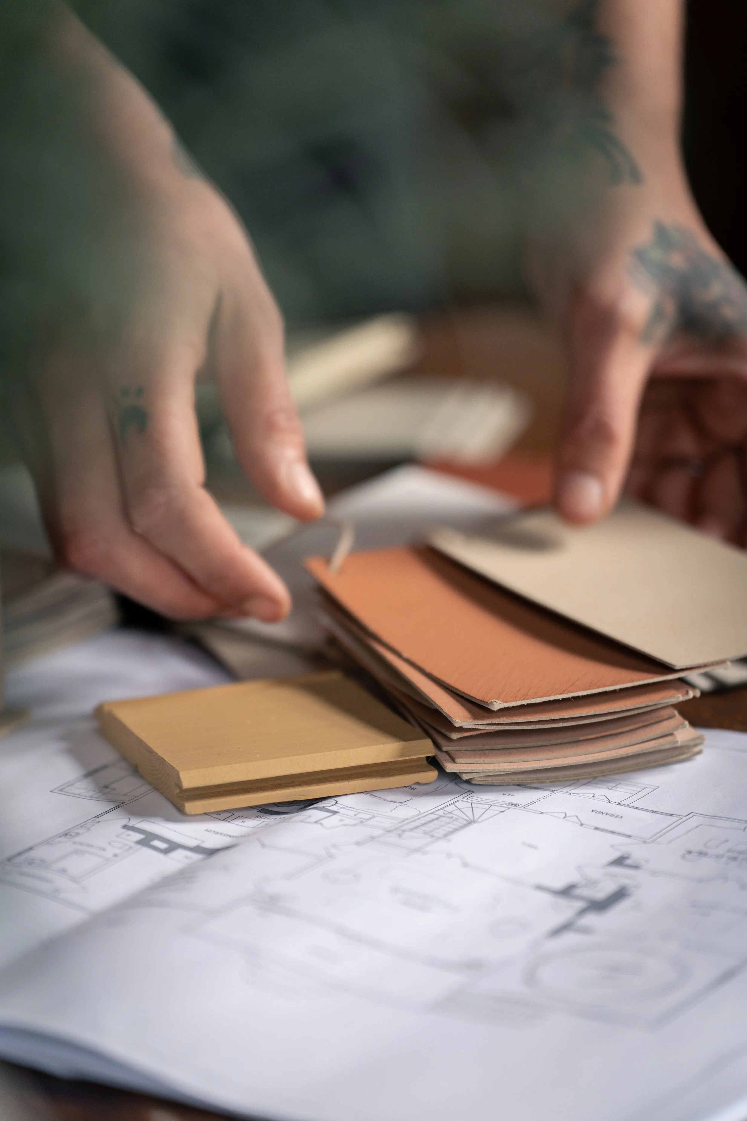 Close-up of tattooed hands flipping through color swatches over architectural blueprints.