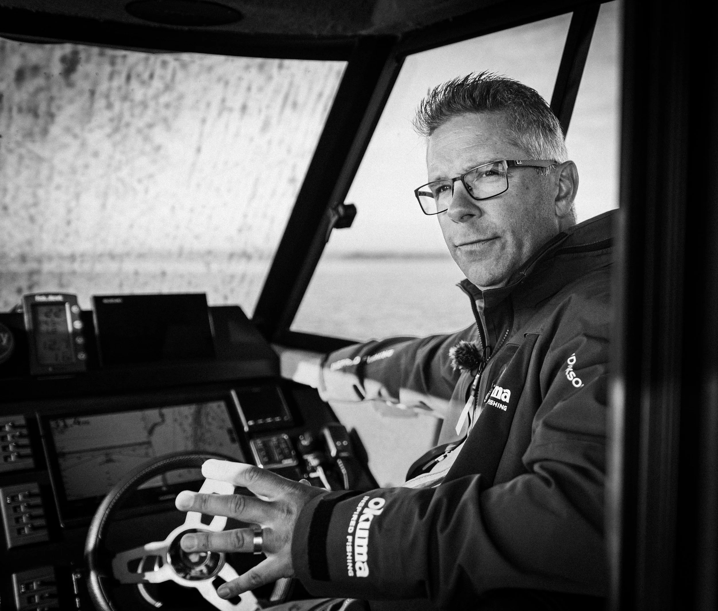A man wearing glasses and a jacket sitting in the cockpit of a boat, looking at the camera with a serious expression.