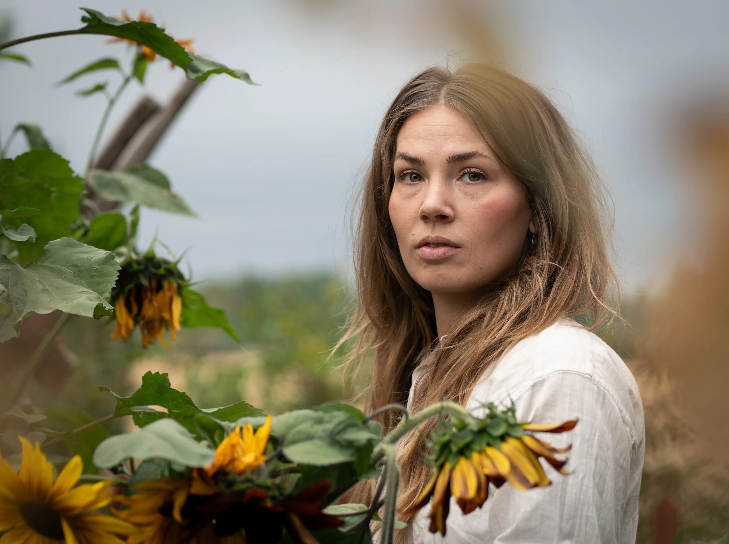 A woman with long, wavy brown hair standing outdoors among sunflowers, wearing a white shirt, with a neutral expression on her face.