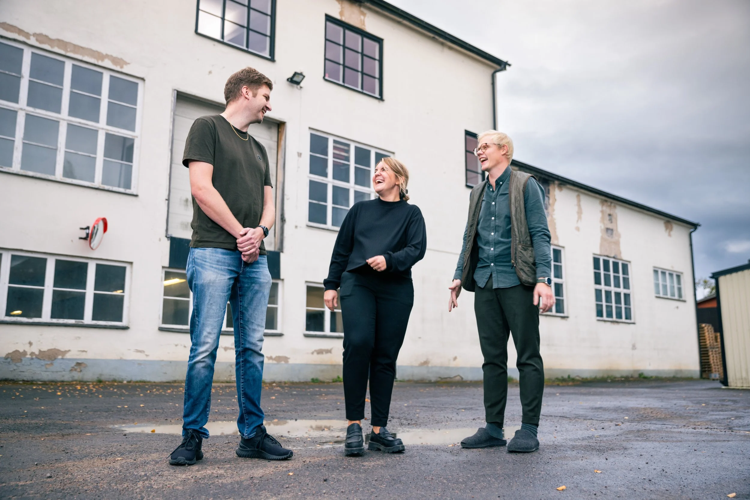 Three young adults, two men and one woman, stand outside and laugh together in front of an old, white building with multiple windows and peeling paint, on a cloudy day.
