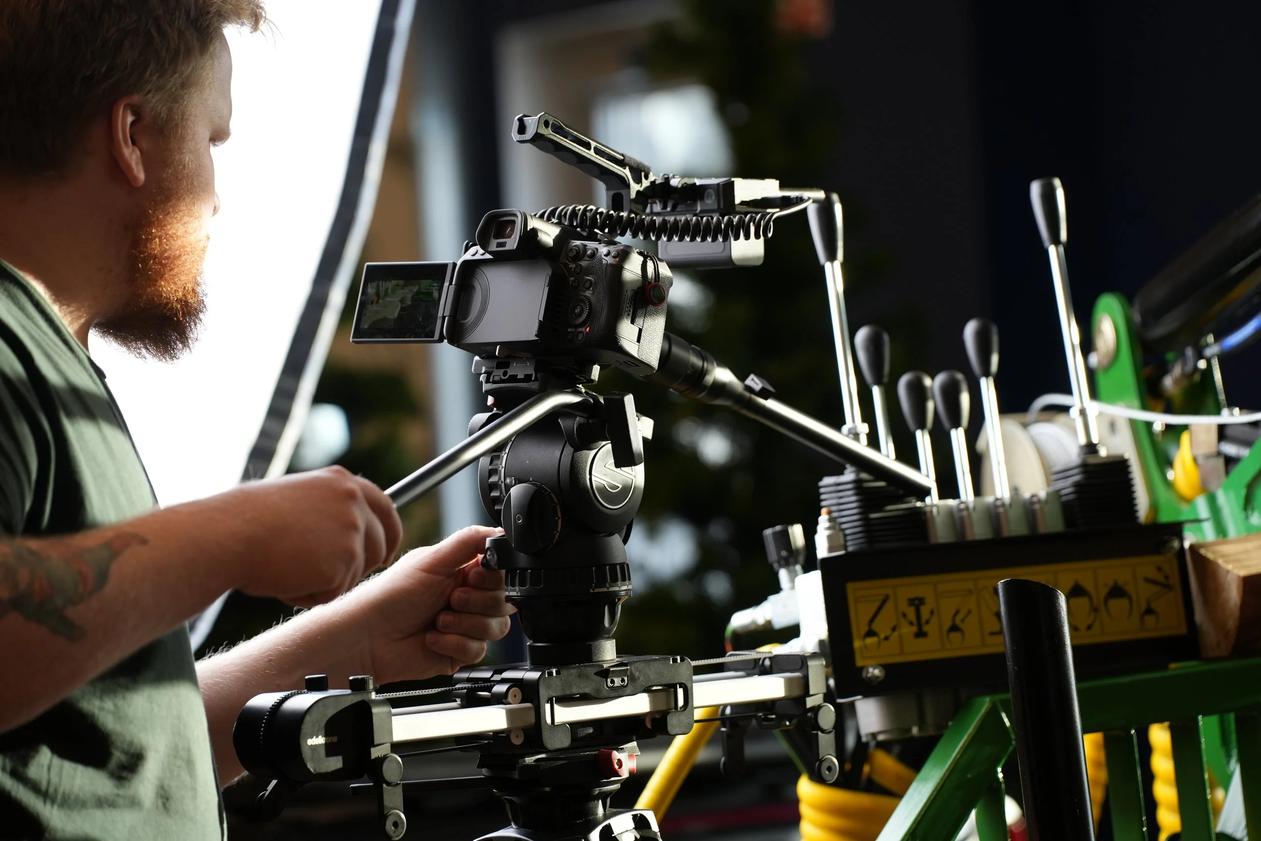 A person operating a camera mounted on a tripod, set up for filming or photography, with various mechanical equipment and tools nearby.
