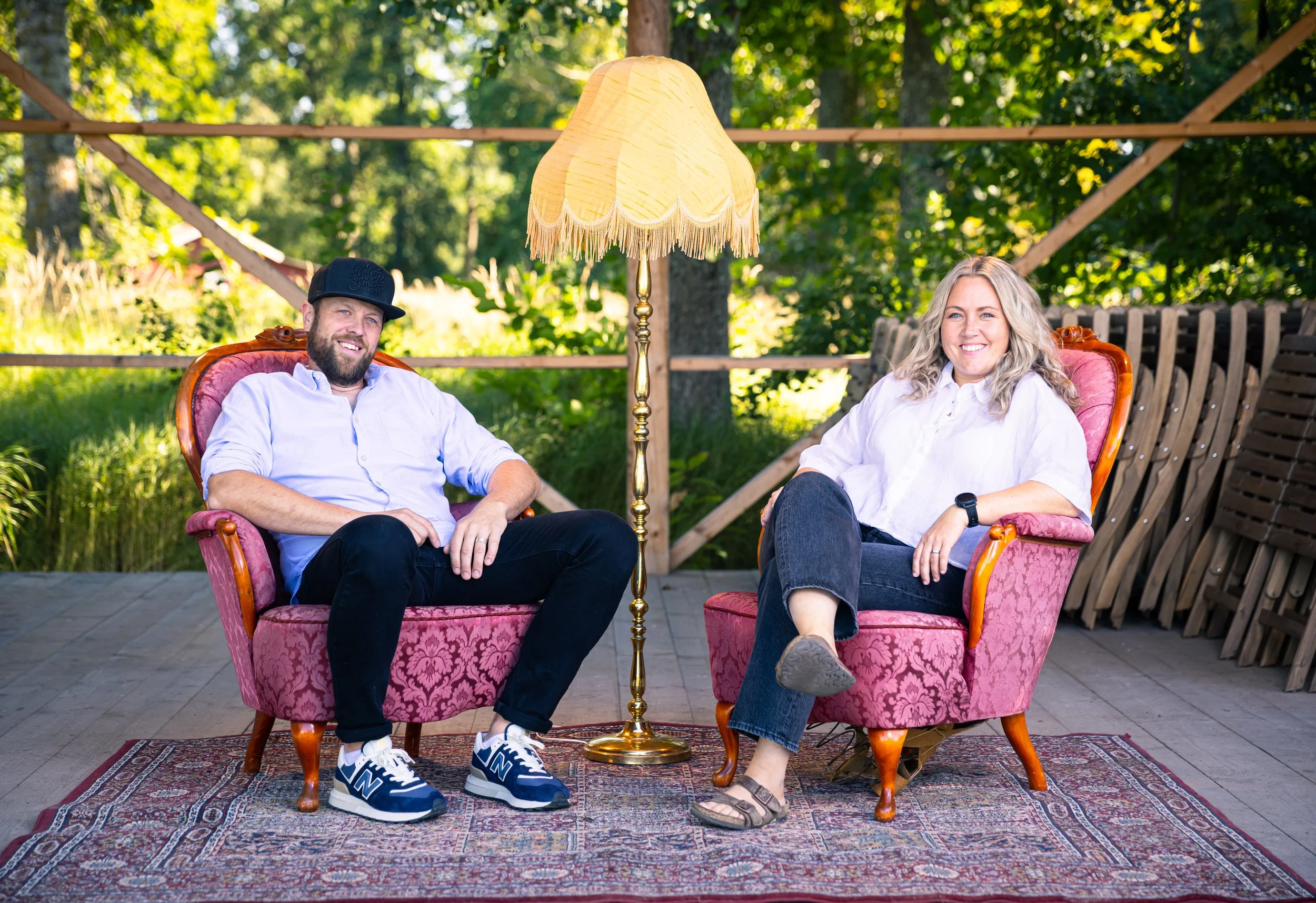 Two people sitting on pink vintage armchairs outdoors with a brass floor lamp between them. The setting is a wooden deck with green trees in the background.