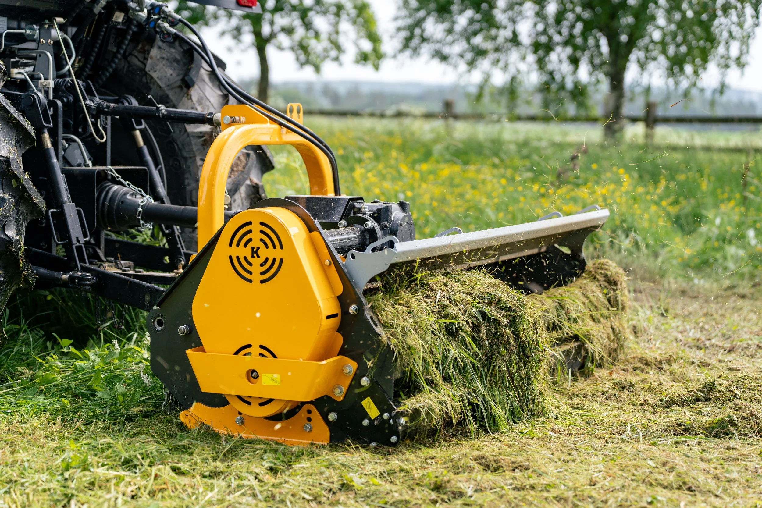 A yellow and black mechanical grass cutter attached to a tractor, trimming grass and weeds in a farm field with trees and a fence in the background.