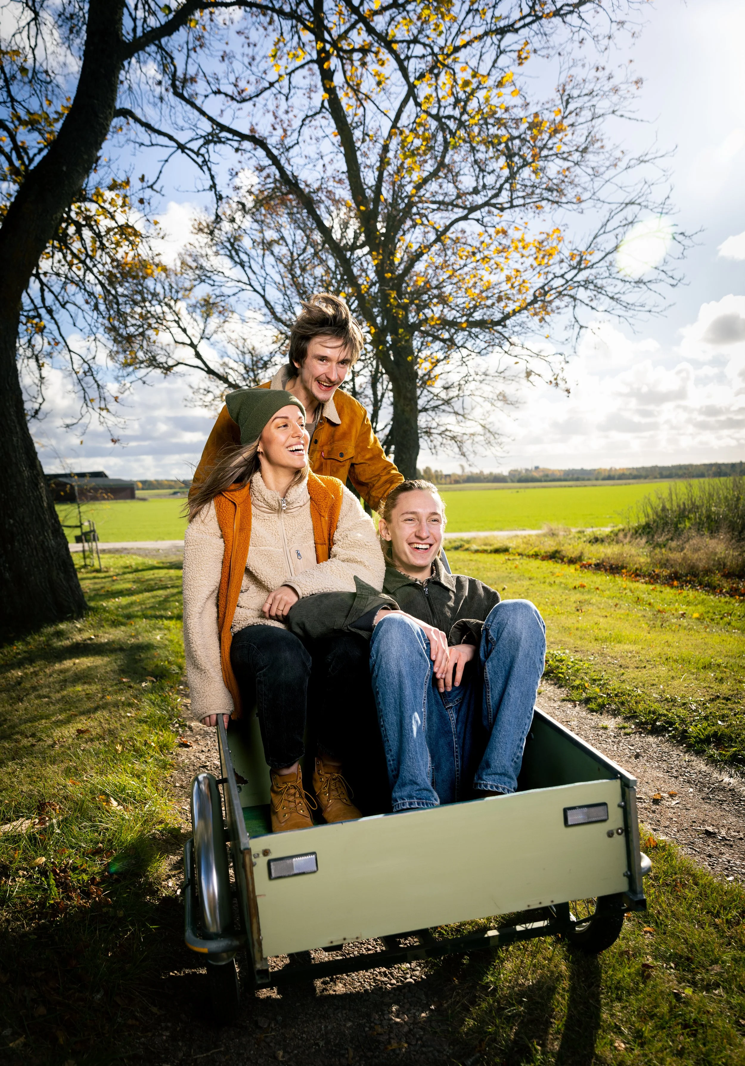 Three young friends enjoying a ride on a small cart in a park on a sunny autumn day with trees and green fields in the background.